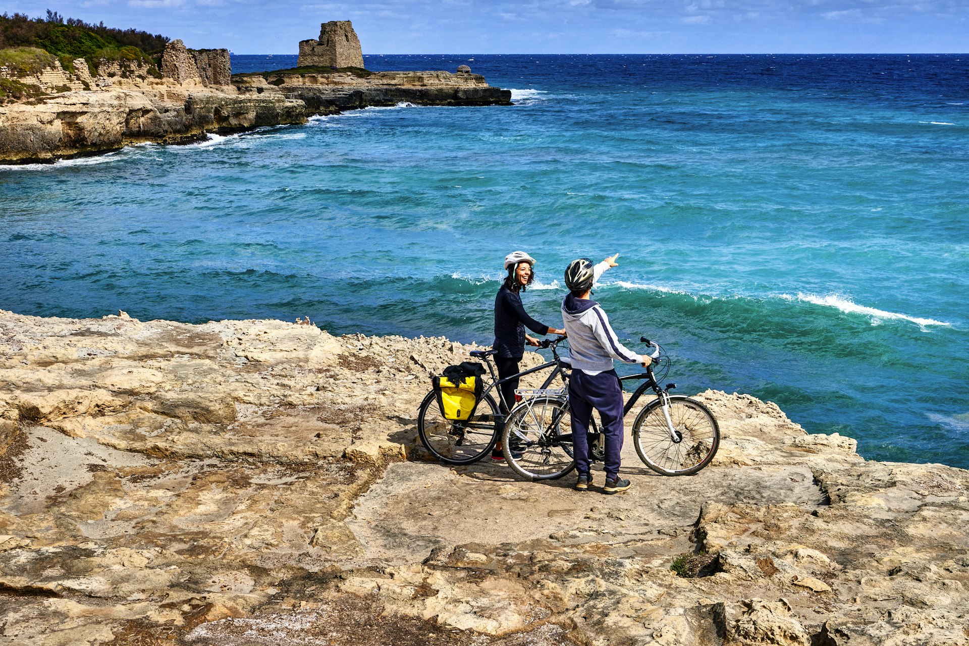 Two people pause with their bicycles on a clifftop looking out over the blue sea and the undulating coastline
