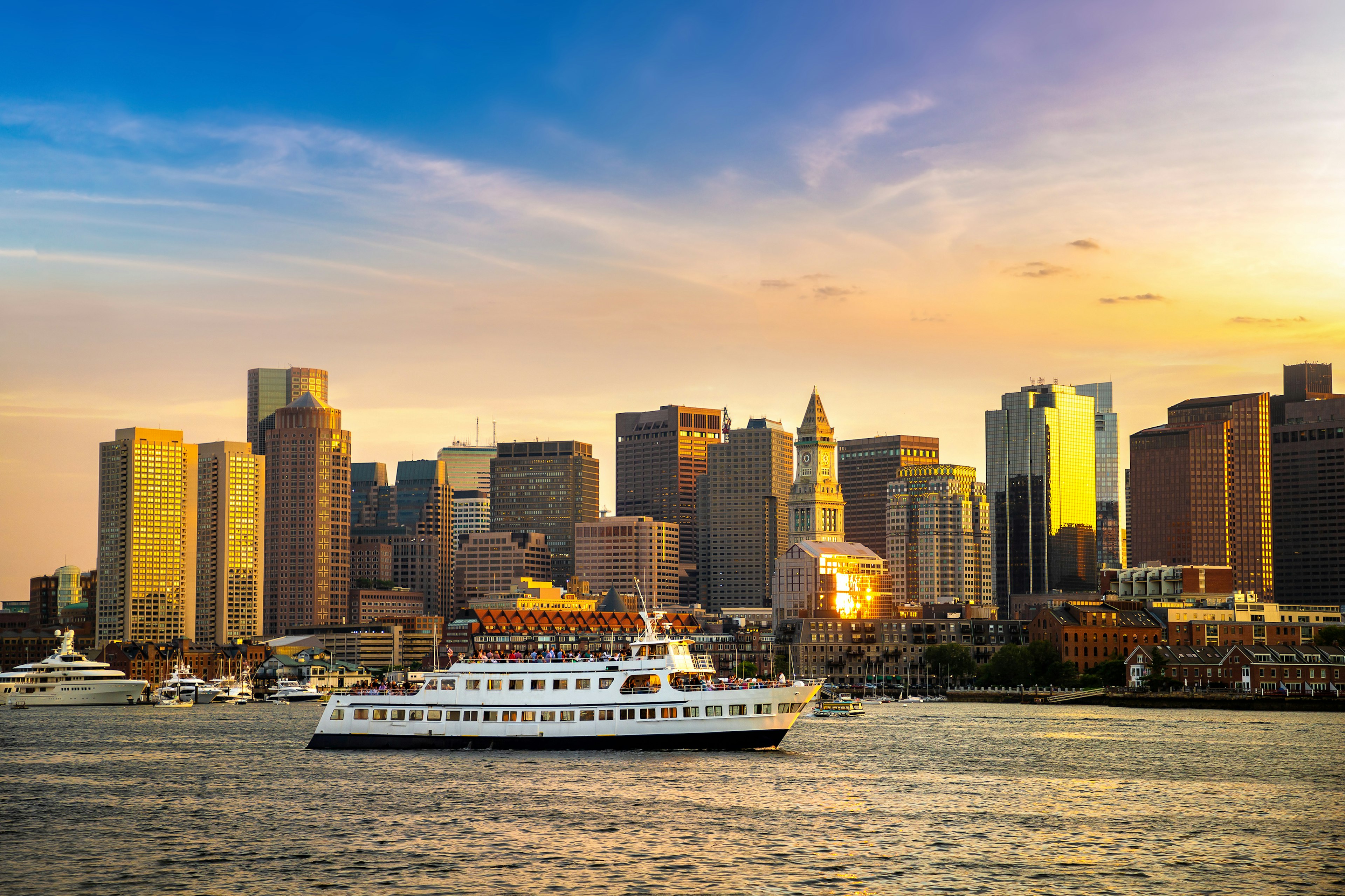 A ferry full of people cruises through Boston Harbor with the sunset reflecting on the tall buildings in the background