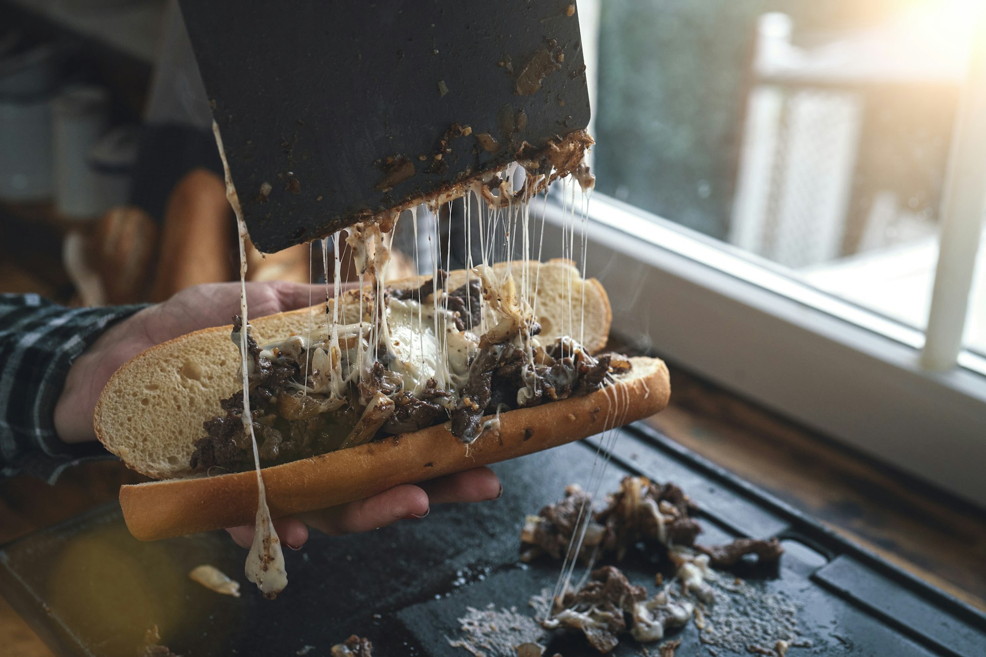 Hot, melted stringy cheese being pulled away from a Philly cheesesteak sandwich in a kitchen