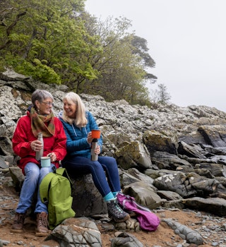 Senior women on a hiking staycation in Dumfries and Galloway, Scotland. They are at the coast on a beach, sitting on rocks while taking a break. They are enjoying hot drinks from travel mugs.
1474788241
