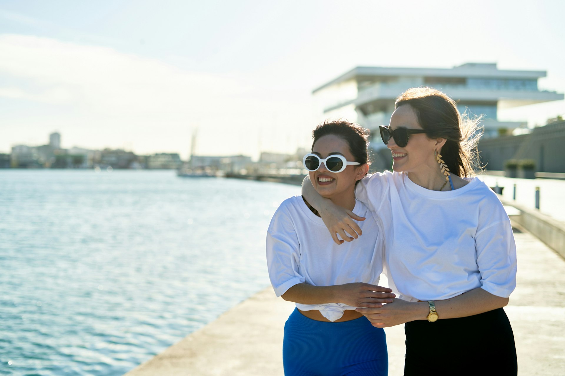Two women walk along a city's waterfront with their arms around each other