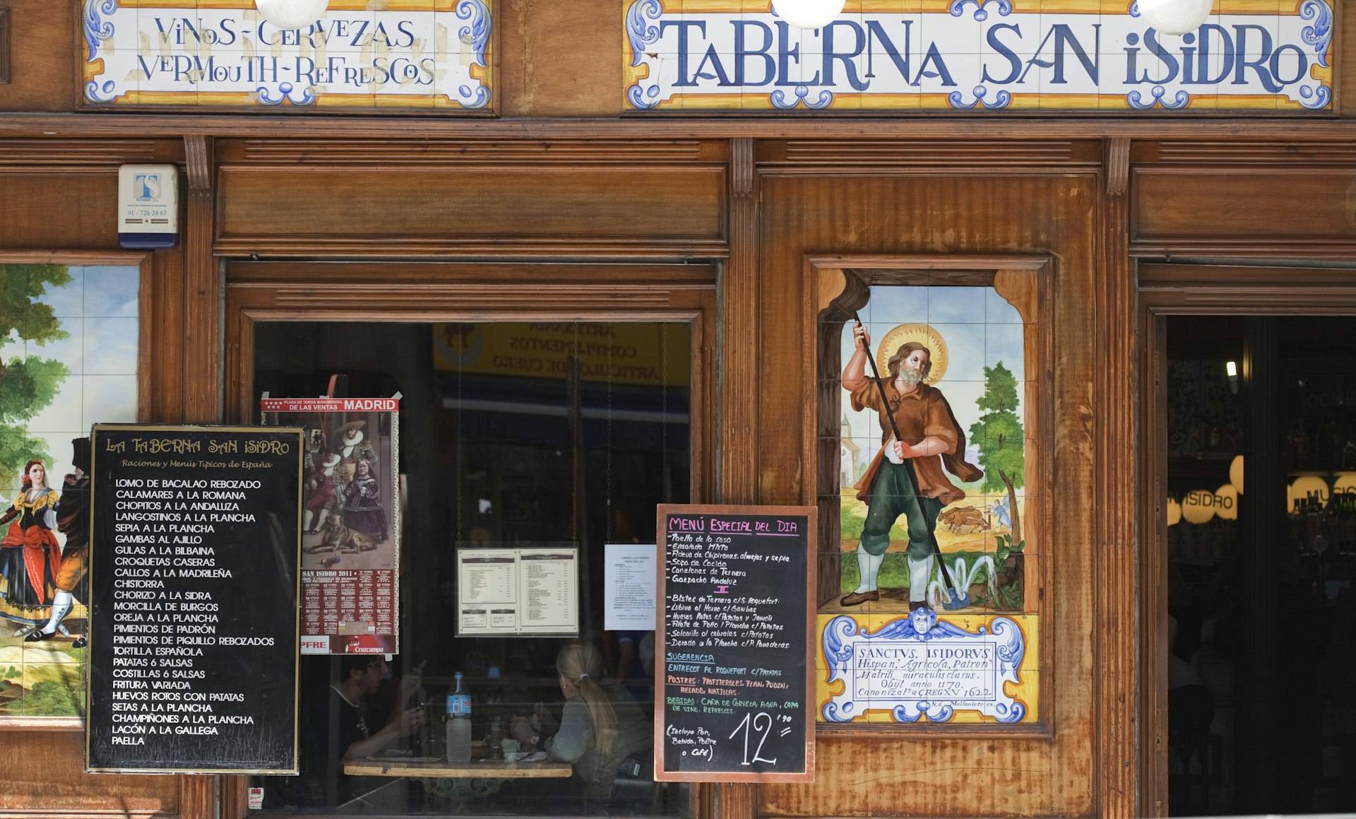 Front view of Taberna de San Isidro, a popular old-style tavern with tile decoration, traditional food and chalkboard menus out front and people seen dining in the front window