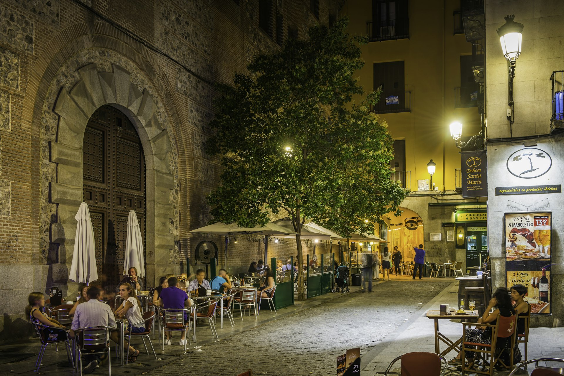 People relaxing at the al fresco tables of a pavement restaurant bar illuminated by the warm lamp light of historic central Madrid, Spain's vibrant capital city.