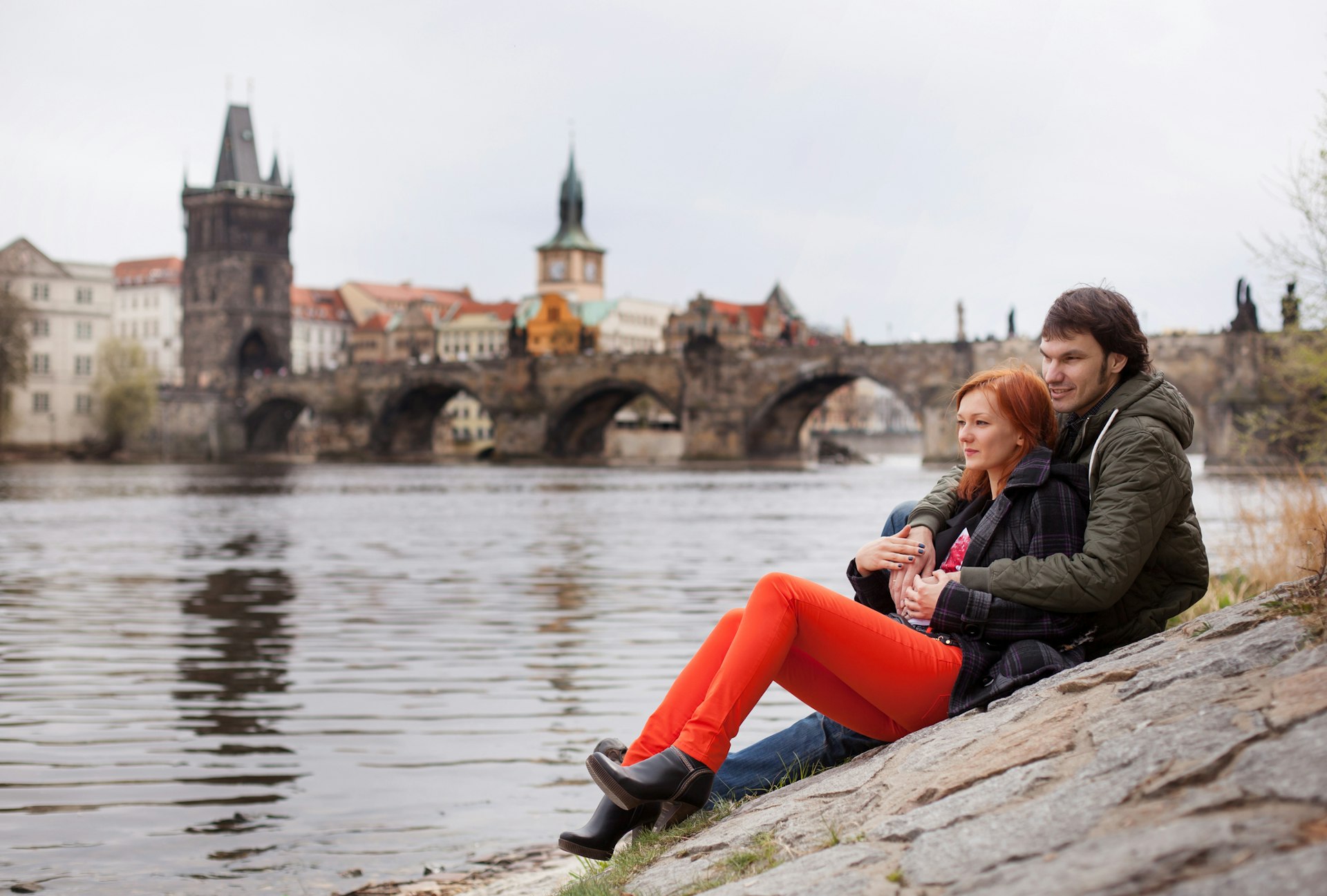 A couple sites by the Vltava River with the Charles Bridge in the background, Prague, Czech Republic
