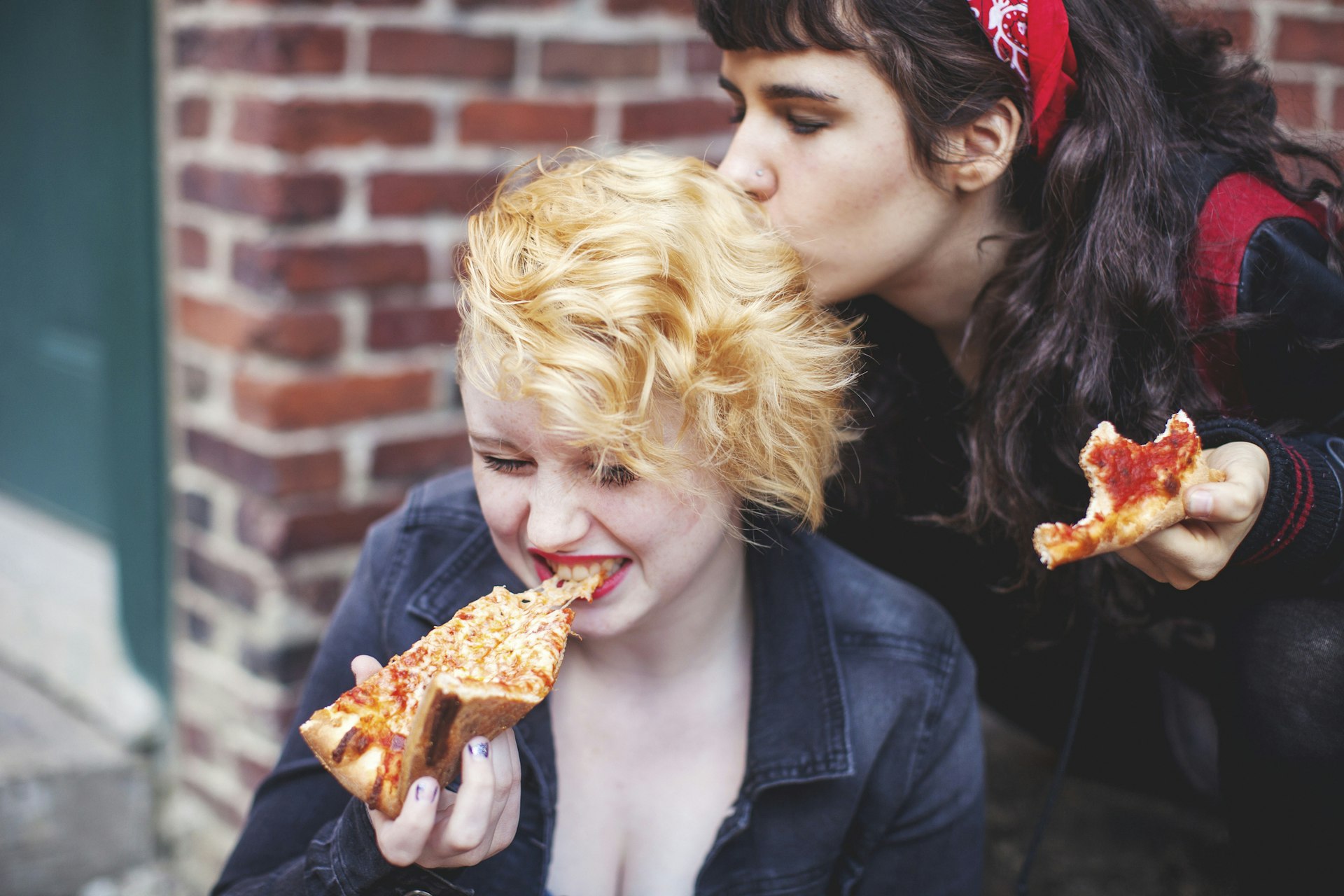 Two women eating pizza on the street in Boston, one is kissing the back of the other's head