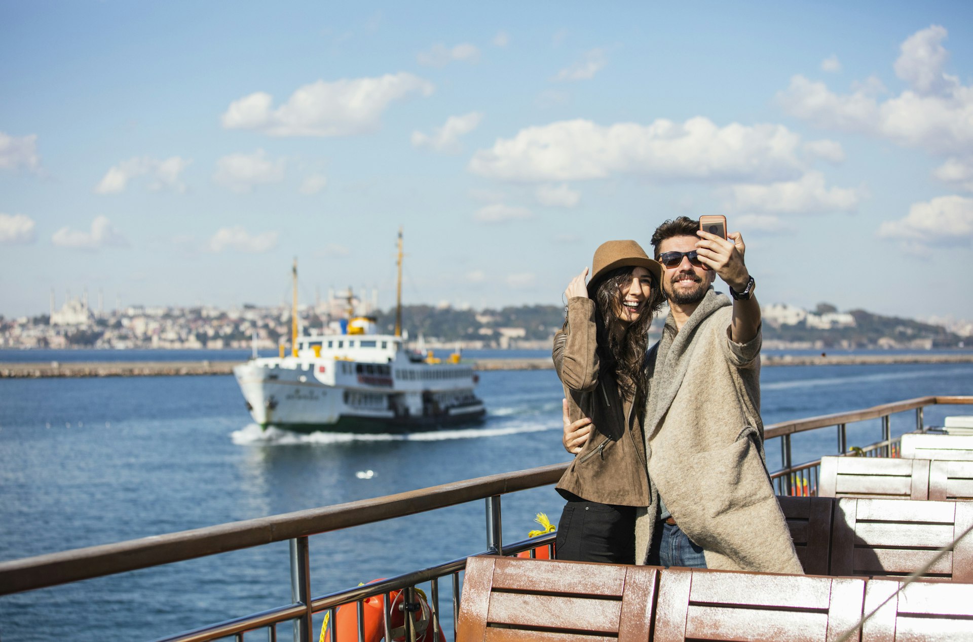 A couple take a smiling selfie as they ride on the ferry with the Istanbul skyline in the background