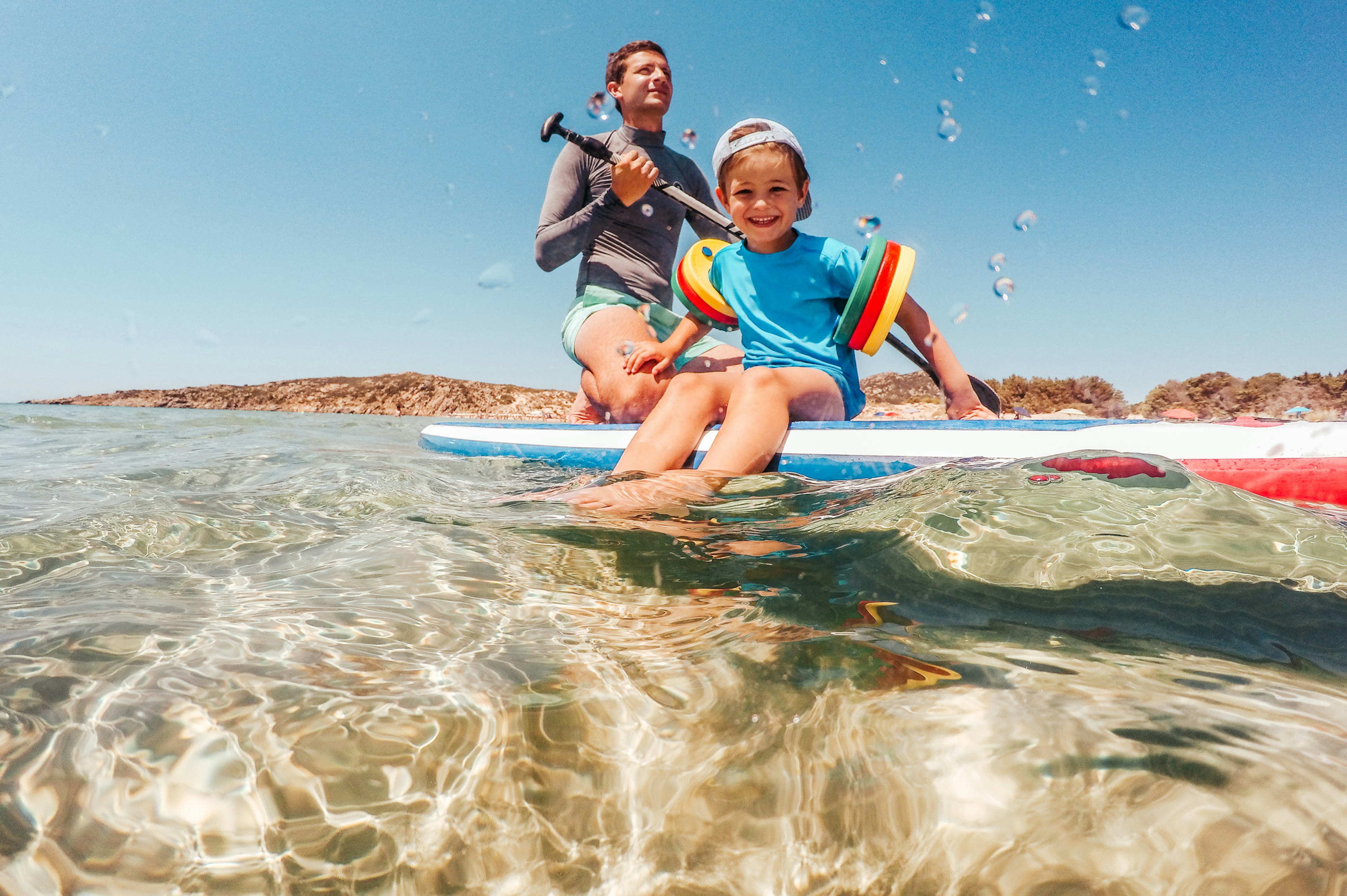 A small boy sat on a paddleboard out at sea smiles at the camera as his dad paddles them along