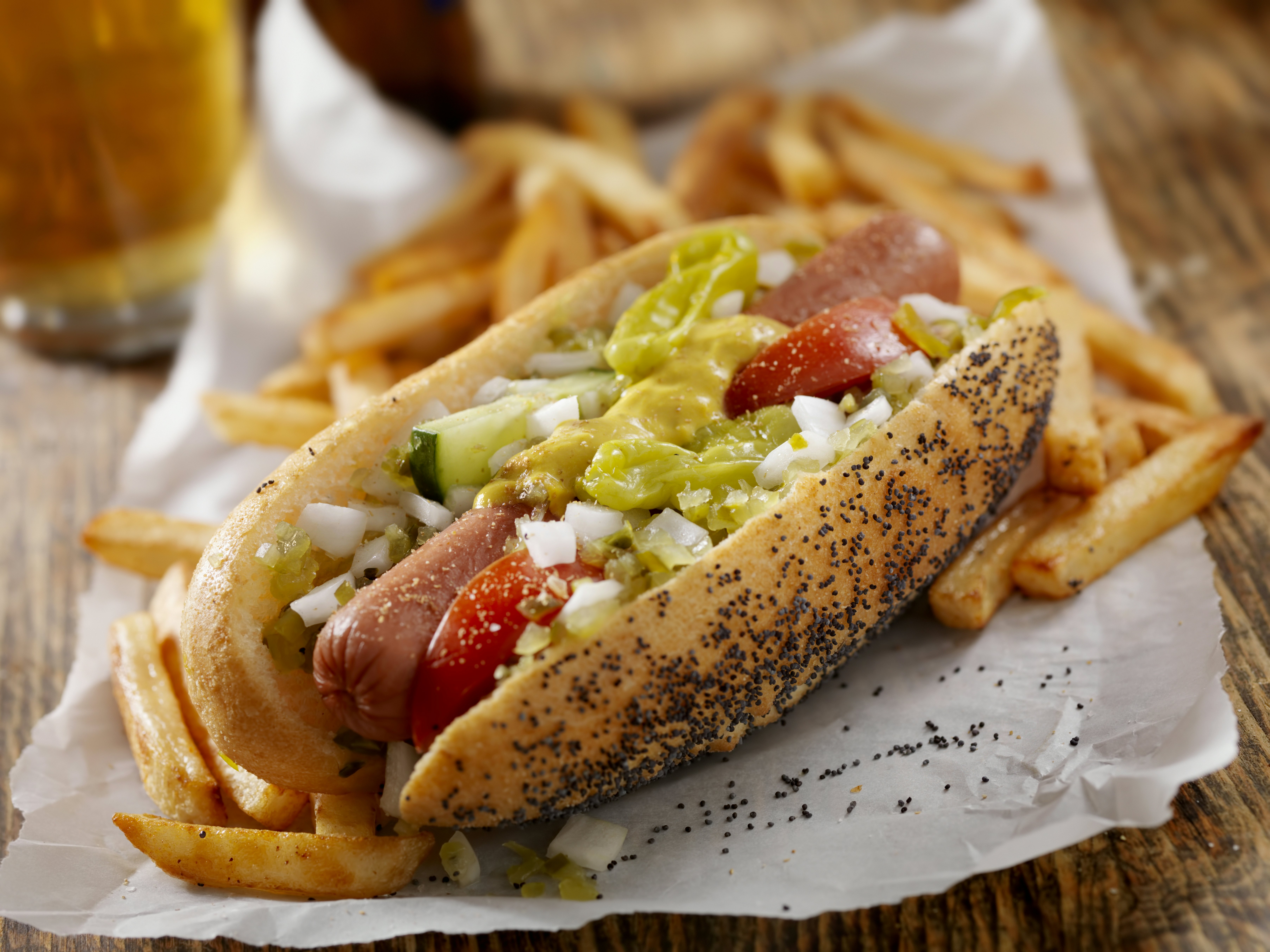 Classic Chicago hot dog served with fries and a beer. The hot dog has fresh tomatoes, diced onions, green relish, peppers, pickle, yellow mustard and a dash of celery salt.