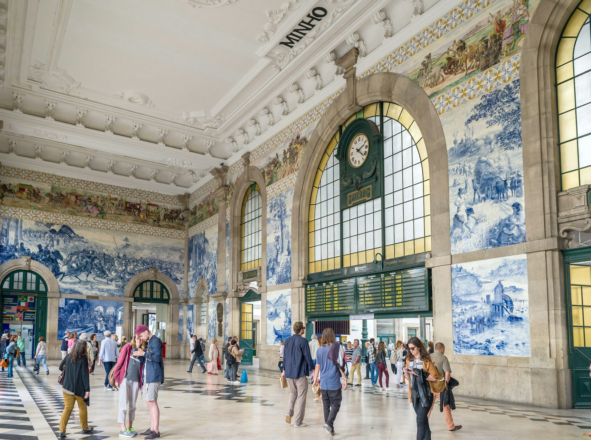 A station concourse busy with people. Many are stopping to look at the intricate blue-and-white tiles that decorate the walls