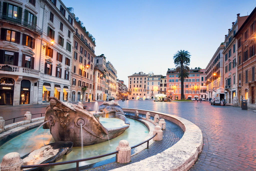 Piazza di Spagna illuminated at dusk, Rome, Italy A low-level sculpture shows a boat sinking in a fountain in a city square