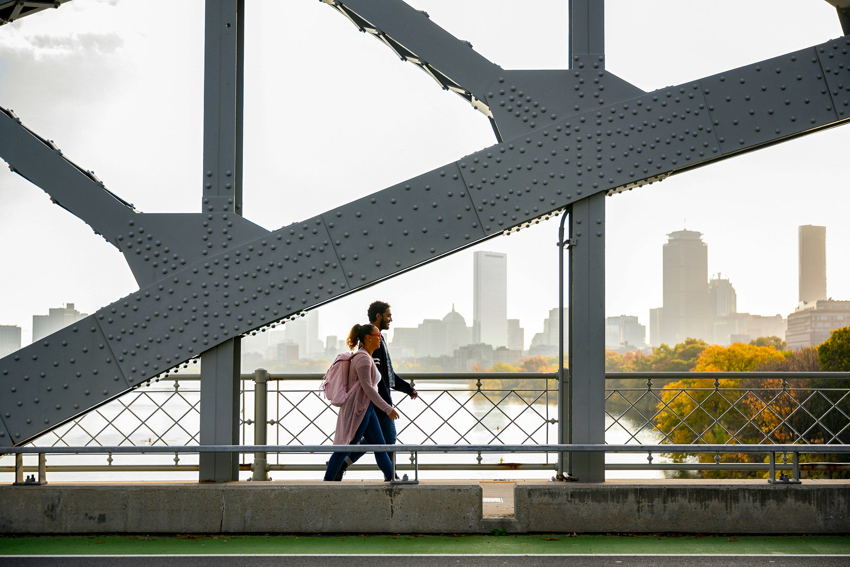 A man and woman walking together along a bridge with the Boston skyline in the background