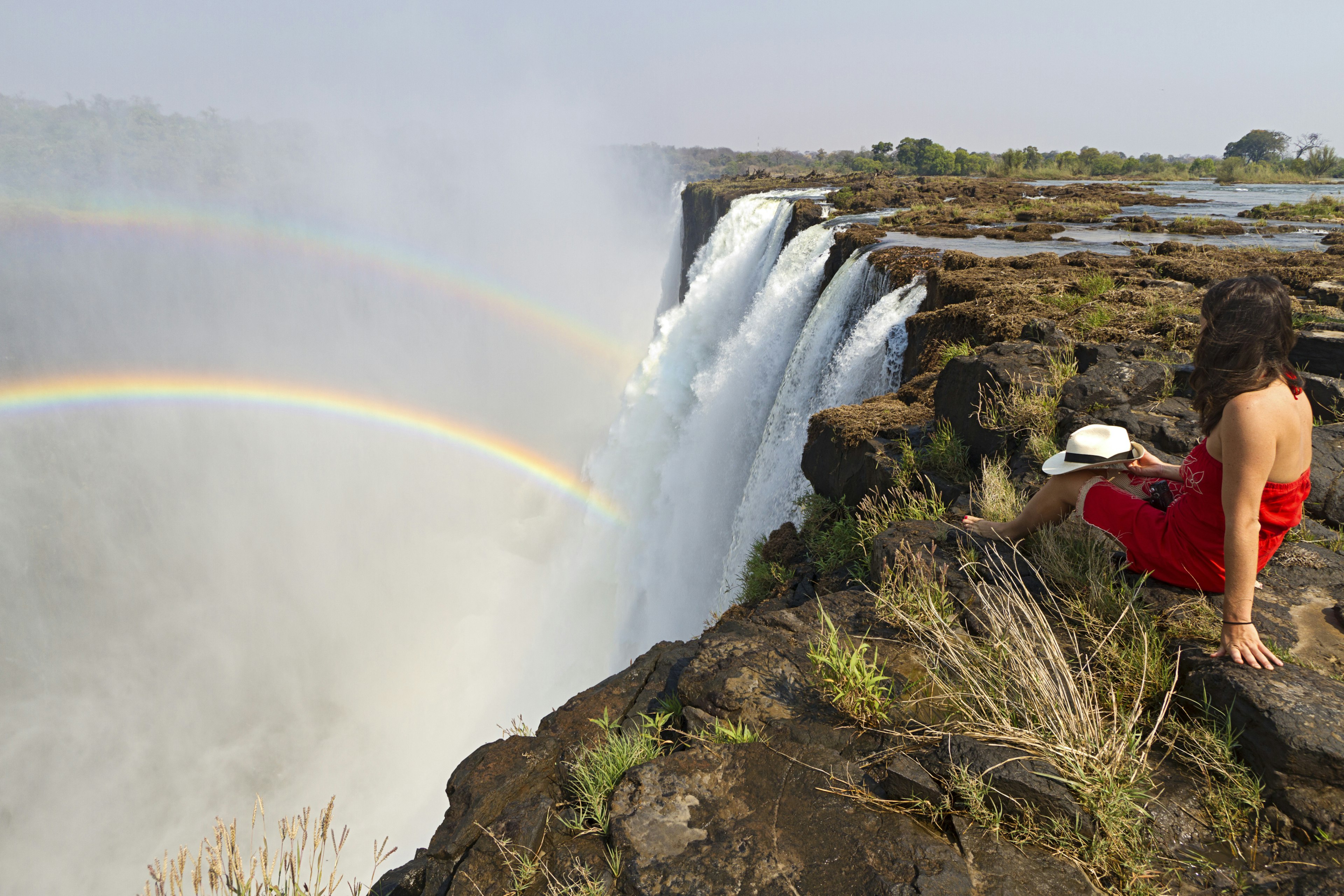 Young woman in a red dress sitting crosslegged at the edge of Victoria Falls which spills out into the gorge below