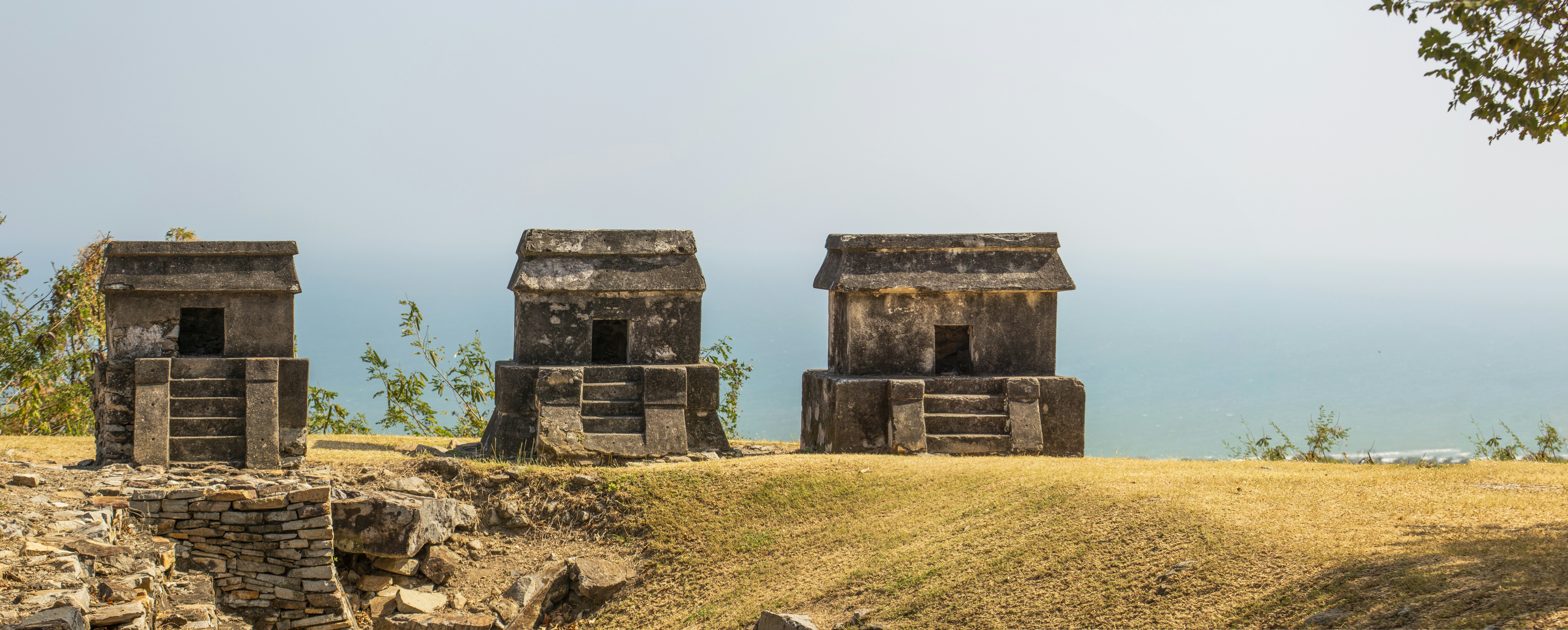 Ancient mausoleum-style tombs on a grassy hillside above the sea on a mostly sunny day.
