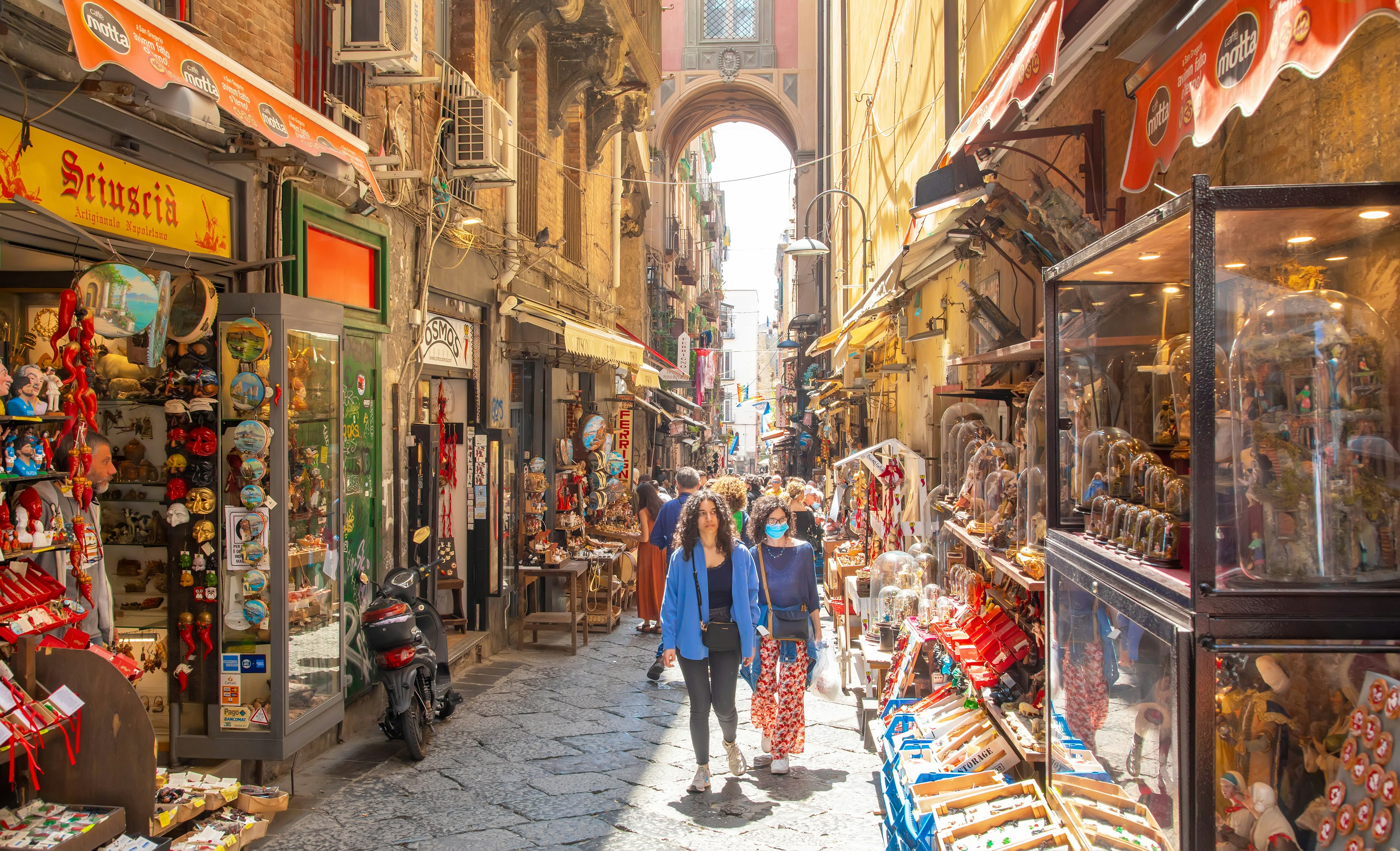 People walk down a narrow alley filled with artisan shops displaying traditional figurines and other crafts in glass cases. A high archway over the street is visible in the background.