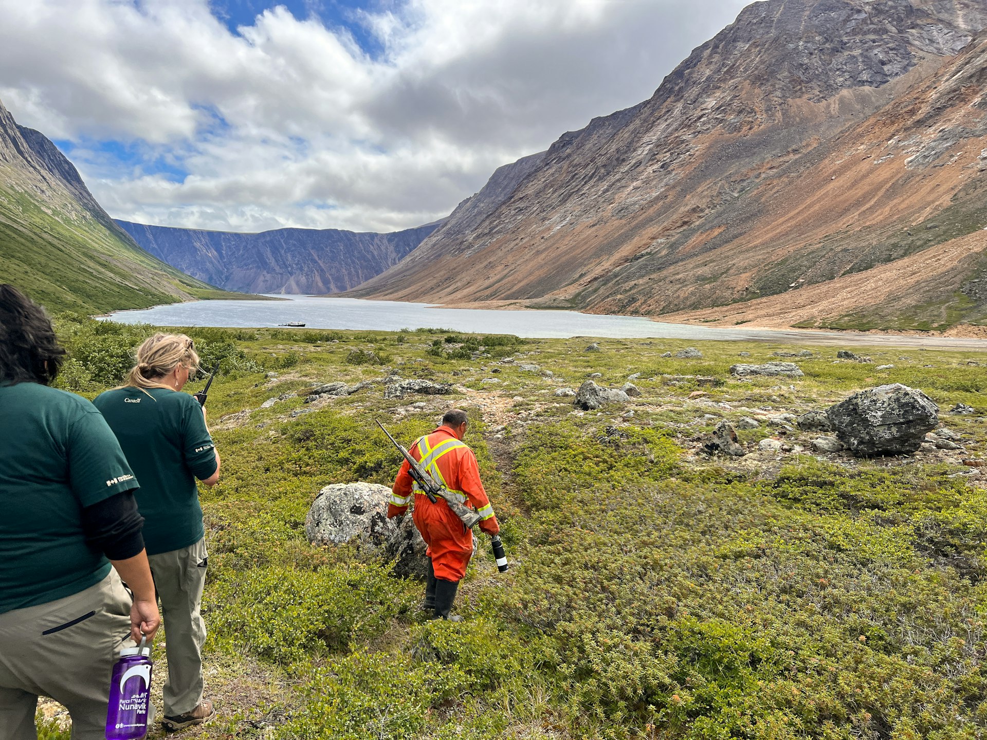 An armed guard wearing high-vis clothing leads a group of people through a mountainous landscape