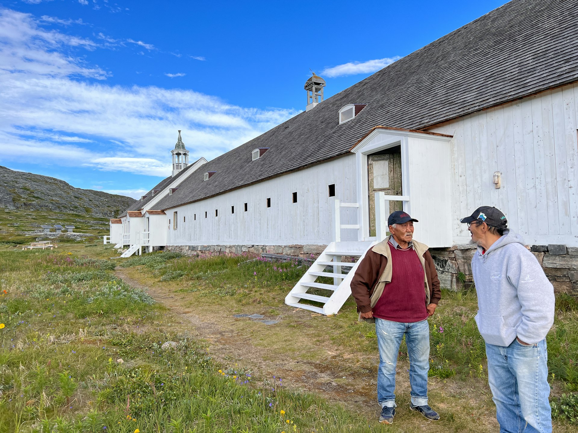 Two men chat with each other outside a long white wooden church building