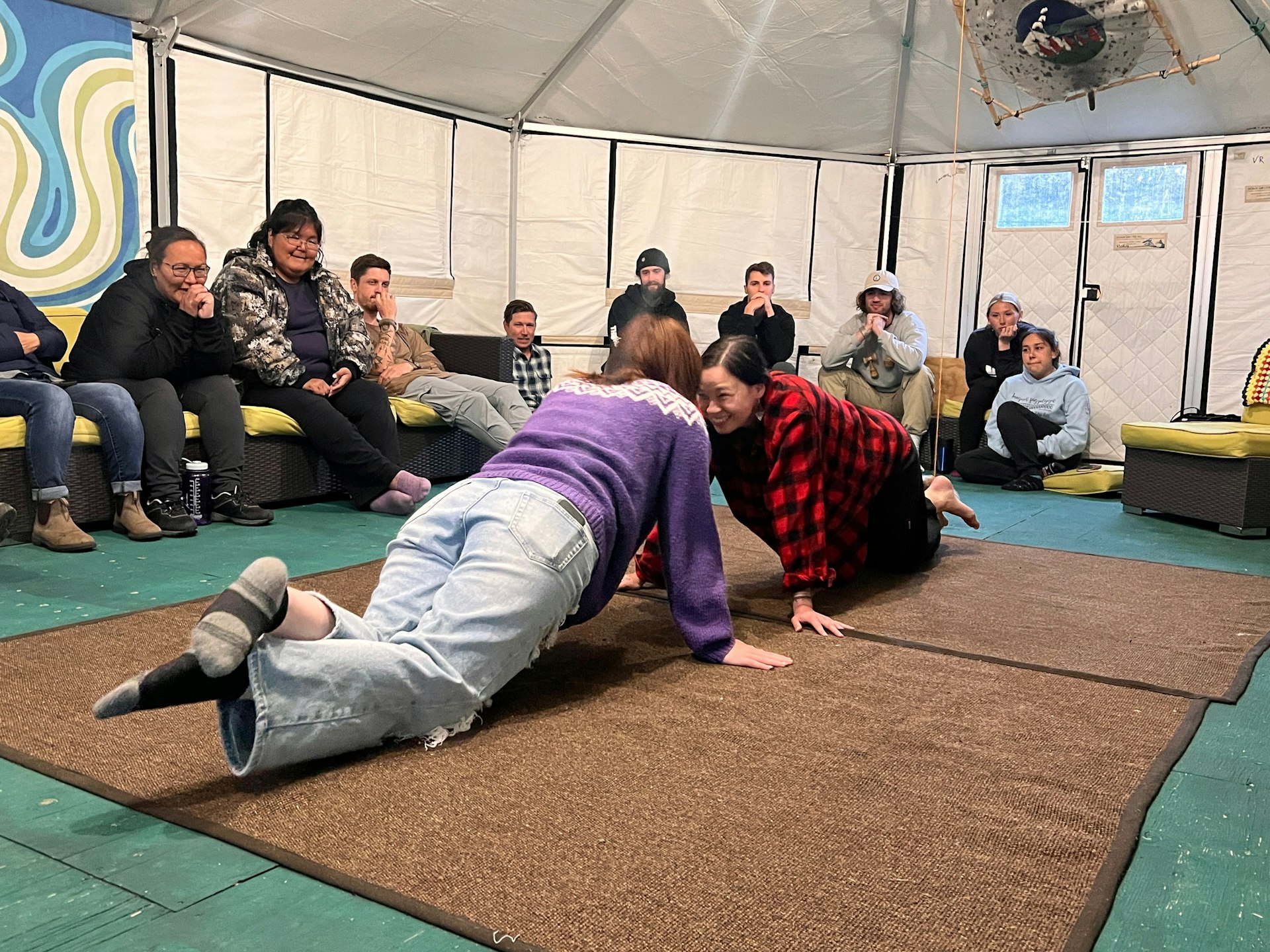 People face each other in a plank position on the floor while other people sit around the edge of the tent watching the games 