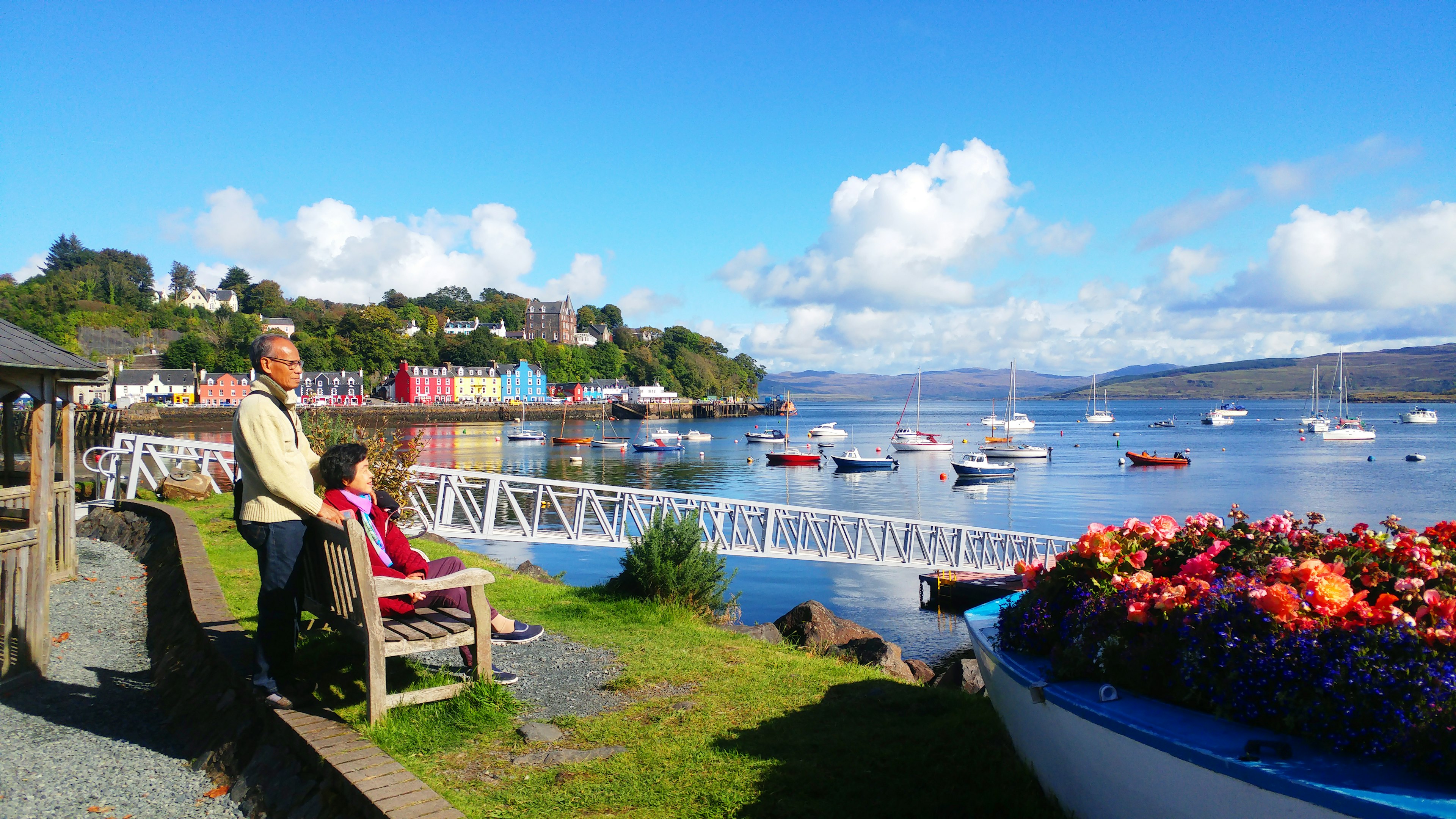 A couple sit on a bench in a harbor town lined with colorful buildings