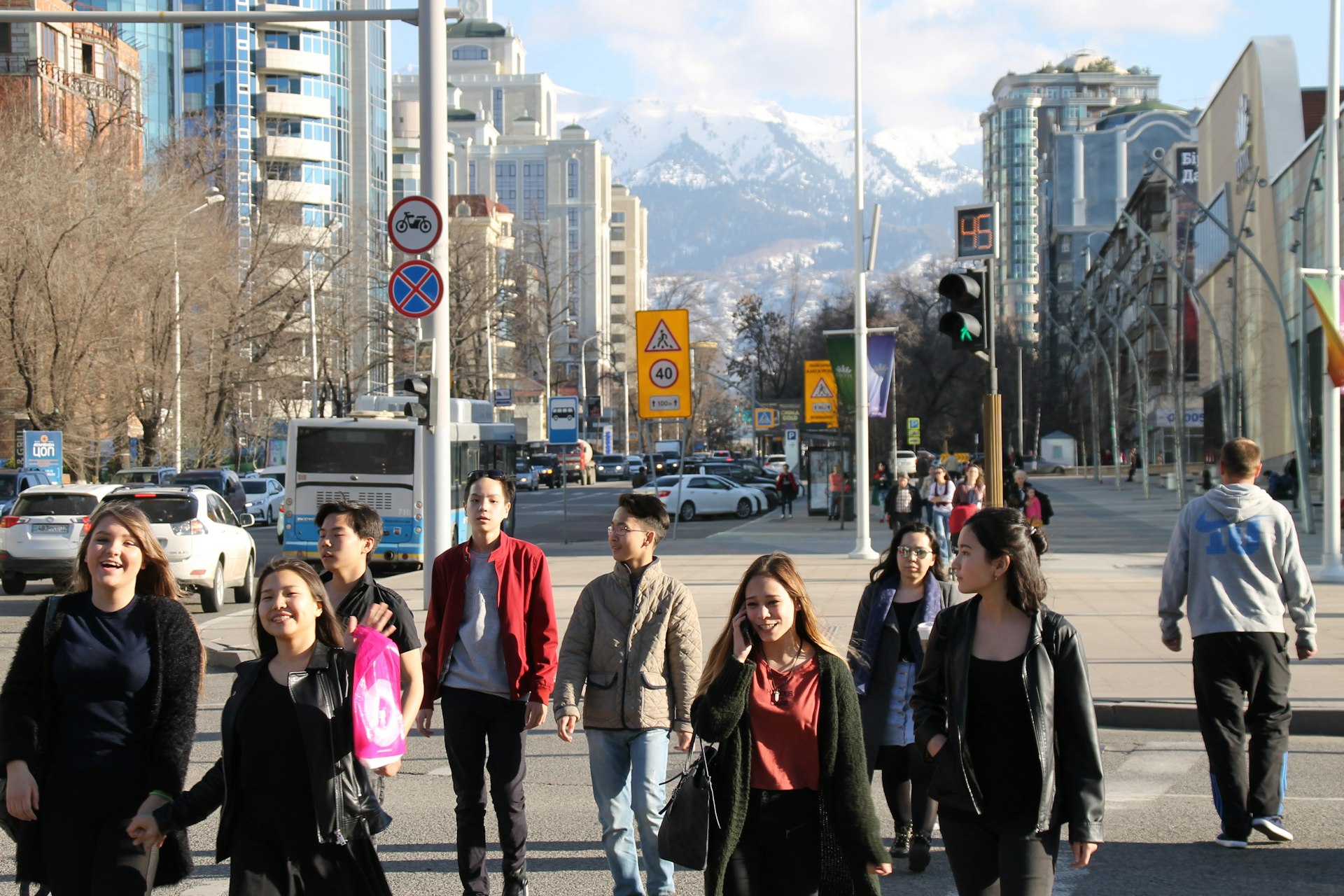 Local Kazakh people walking in the streets among tall buildings with a view of snow-capped peaks in the distance, Almaty, Kazakhstan