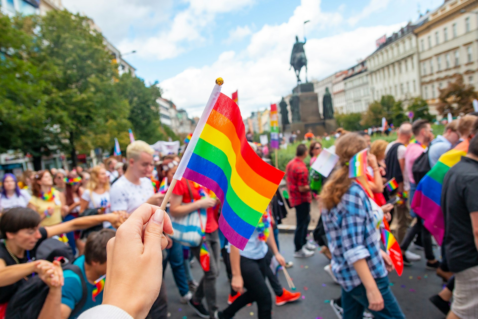 A hand holds up a rainbow flag as a parade of people walk past a large statue of a man on horseback