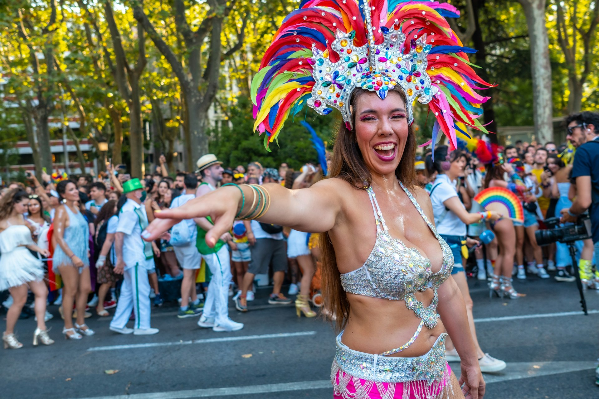 A dancer in a parade wearing a rainbow headdress smiles and points at the camera Title: Pride, Madrid