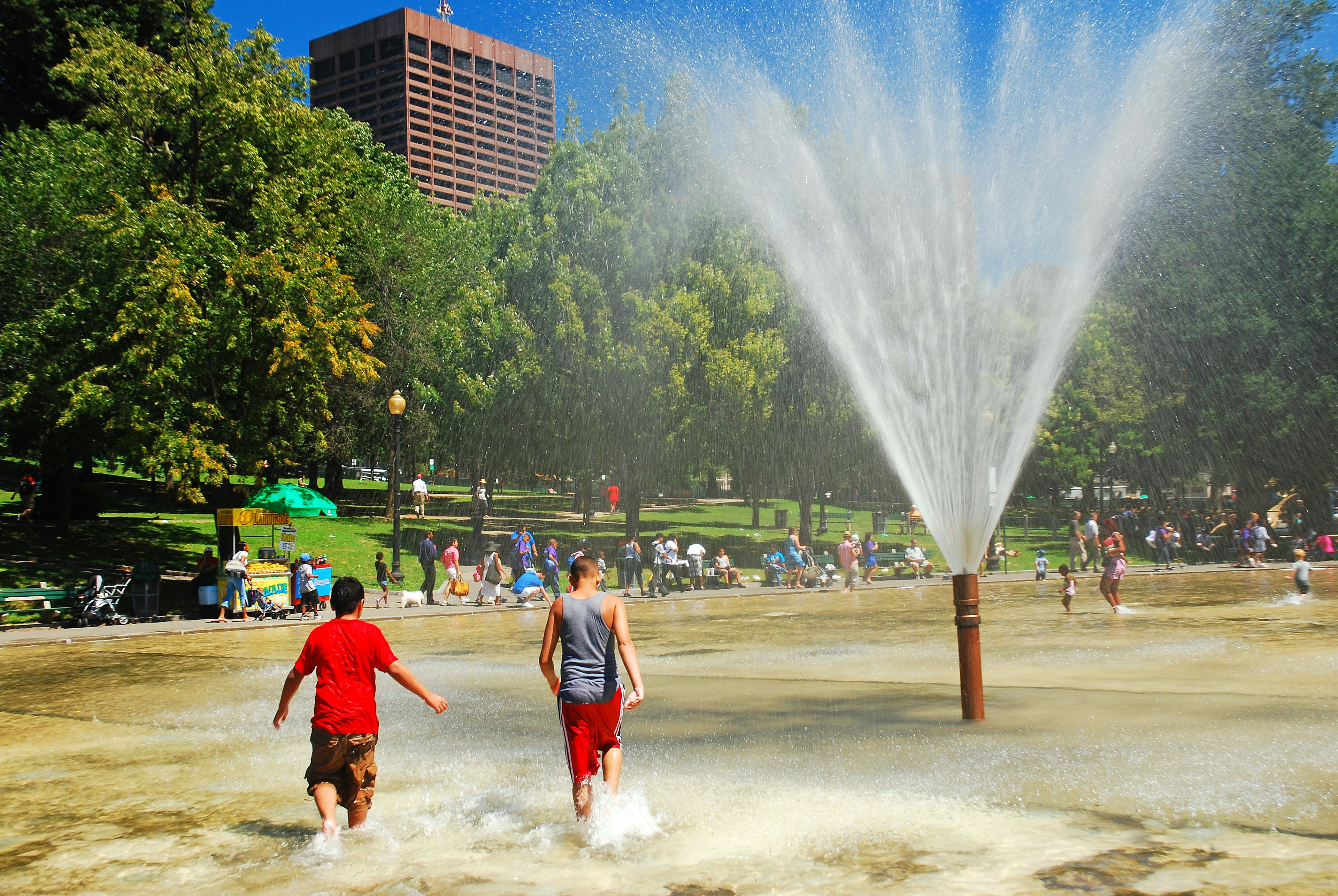 Two teens wander through a shallow outdoor pool with a large jet of water gushing up into the sky on a sunny day