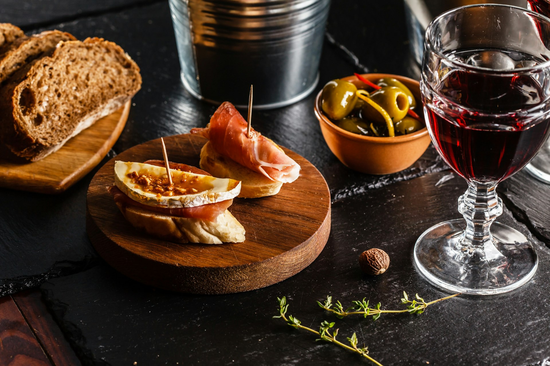 Closeup on a table with a plate of tapas (two slices of bread, one topped with ham and cheese and the other with just ham, held together with toothpicks), slices of plain bread, a dish of olives, sprigs of thyme and a glass of wine