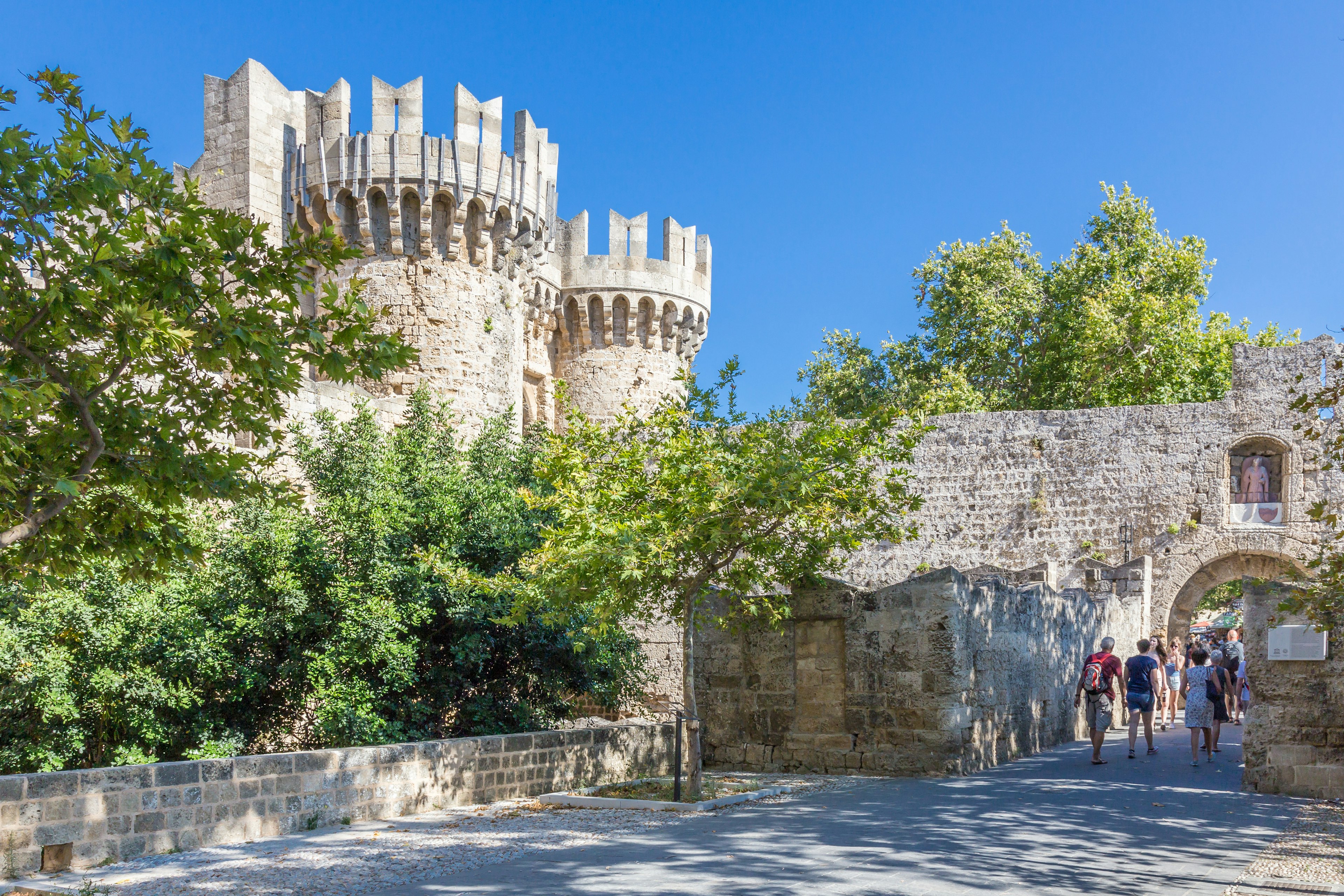 A fortress with two large turrets surrounded by green trees. Visitors stroll through an arched doorway in an old city wall.