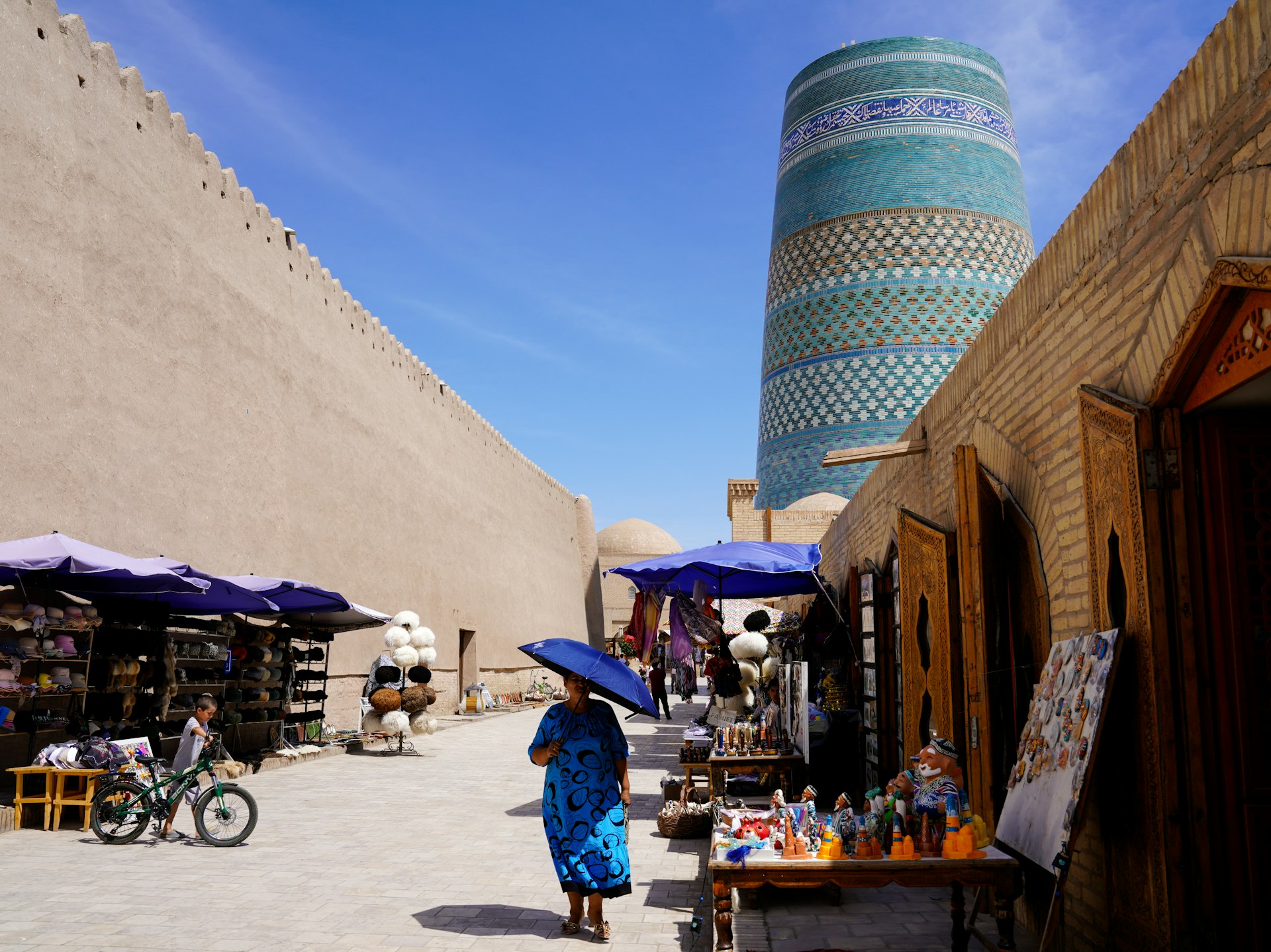 A woman walks by shops with a minaret in the distance, Khiva, Uzbekistan