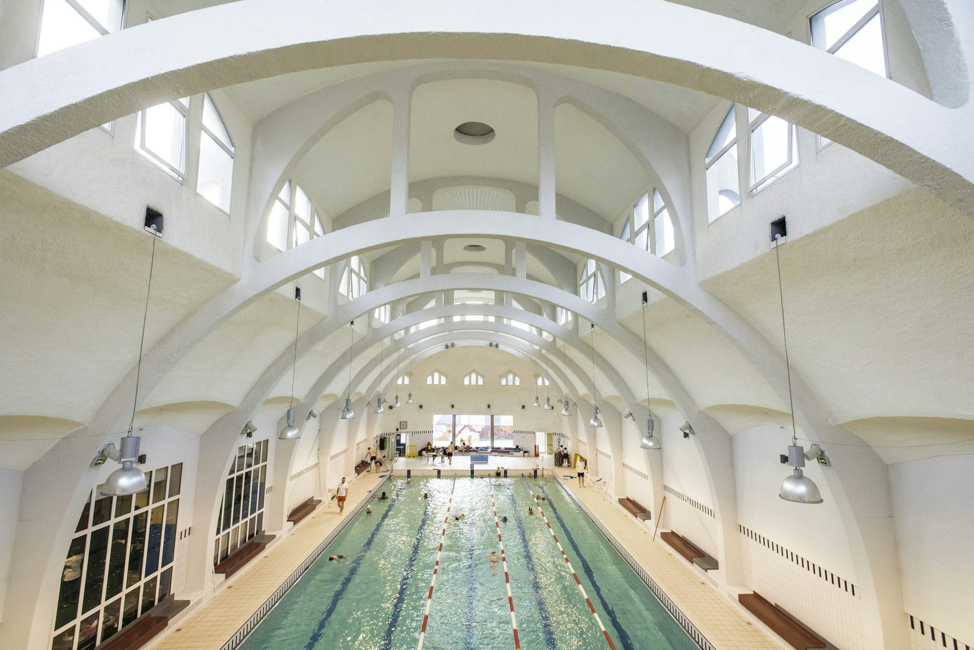 Swimmers swim back and forth in an indoor lap pool beneath white arched ceilings