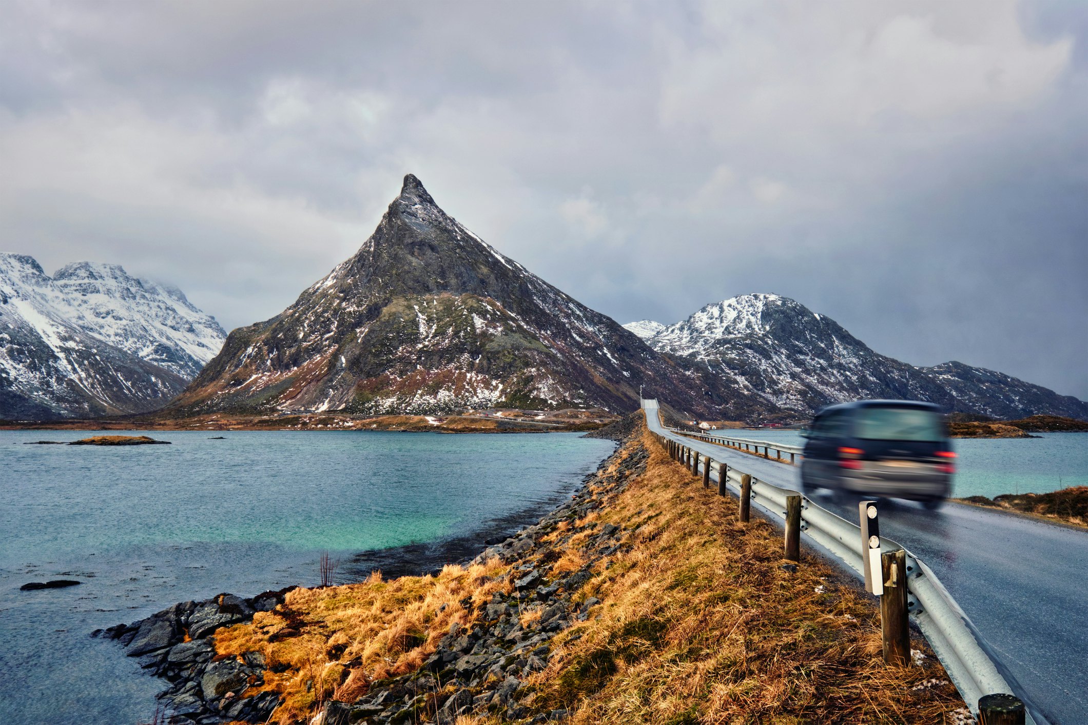 A car heads along a narrow road leading to a bridge across a fjord with a mountain in the distance