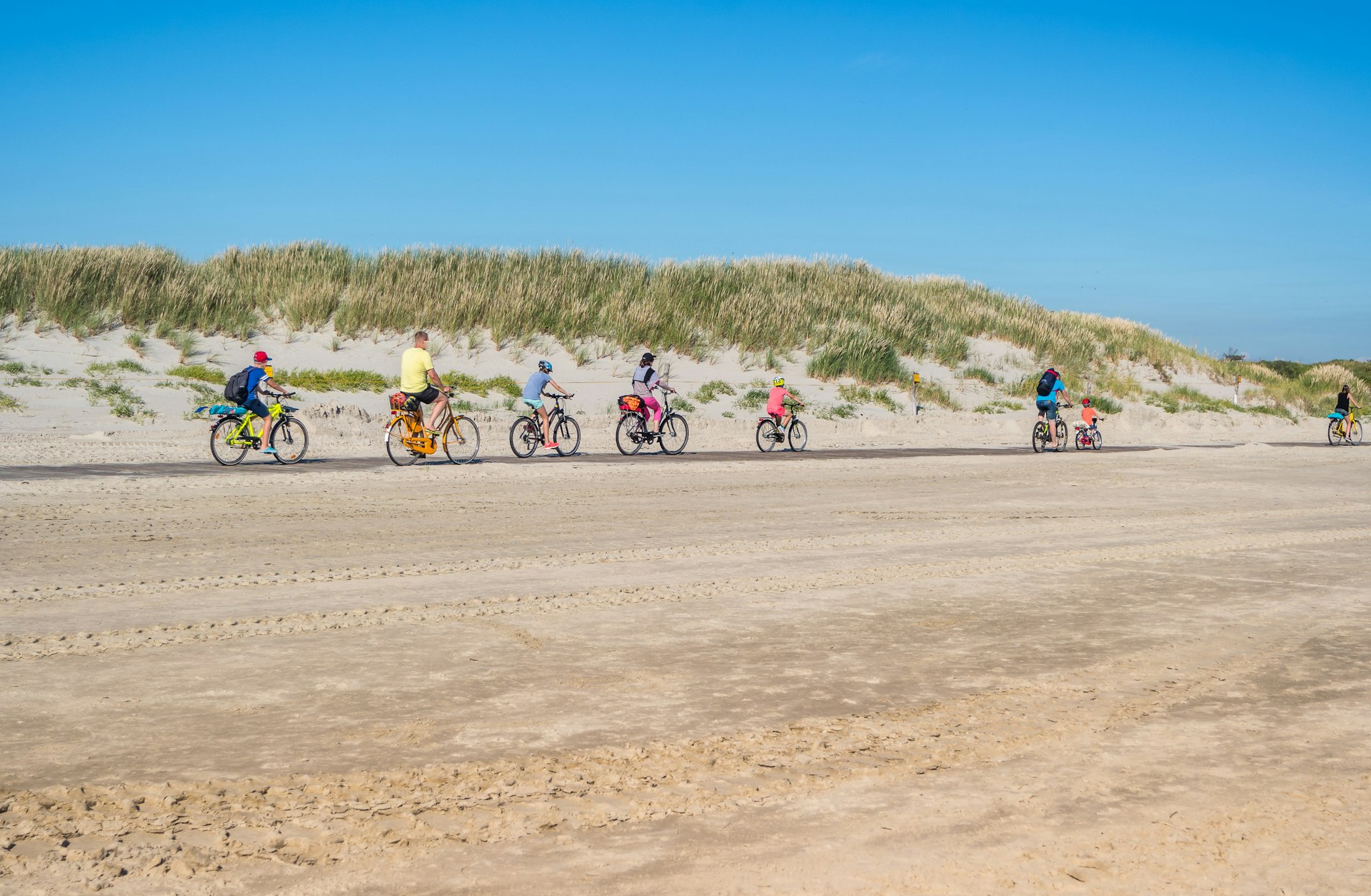 A line of cyclists of all ages follow a bike path along a wide sandy beach