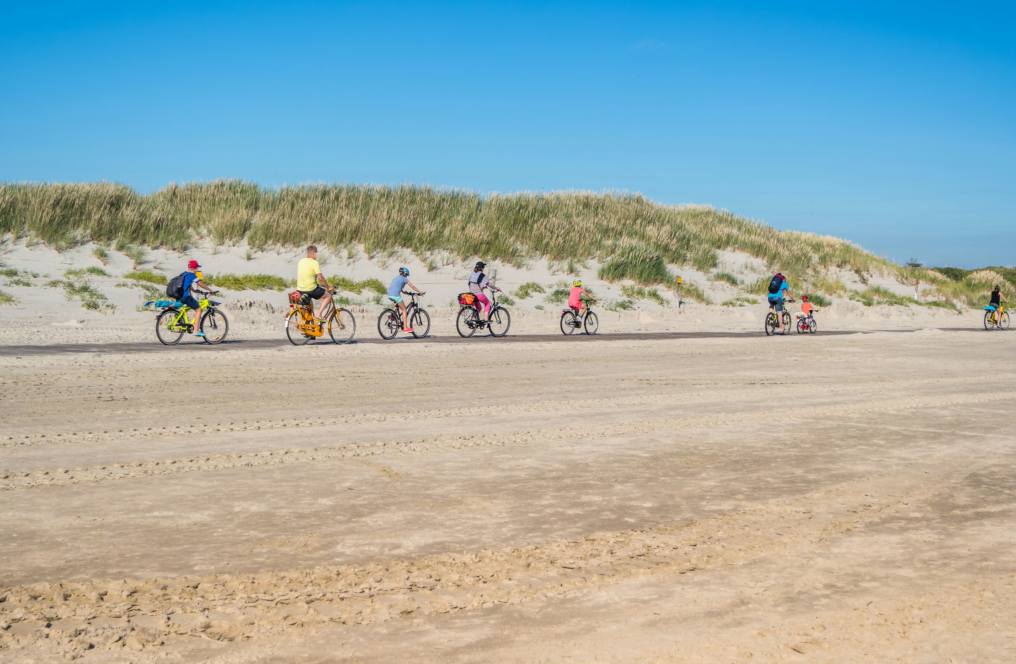 A line of cyclists of all ages follow a bike path along a wide sandy beach