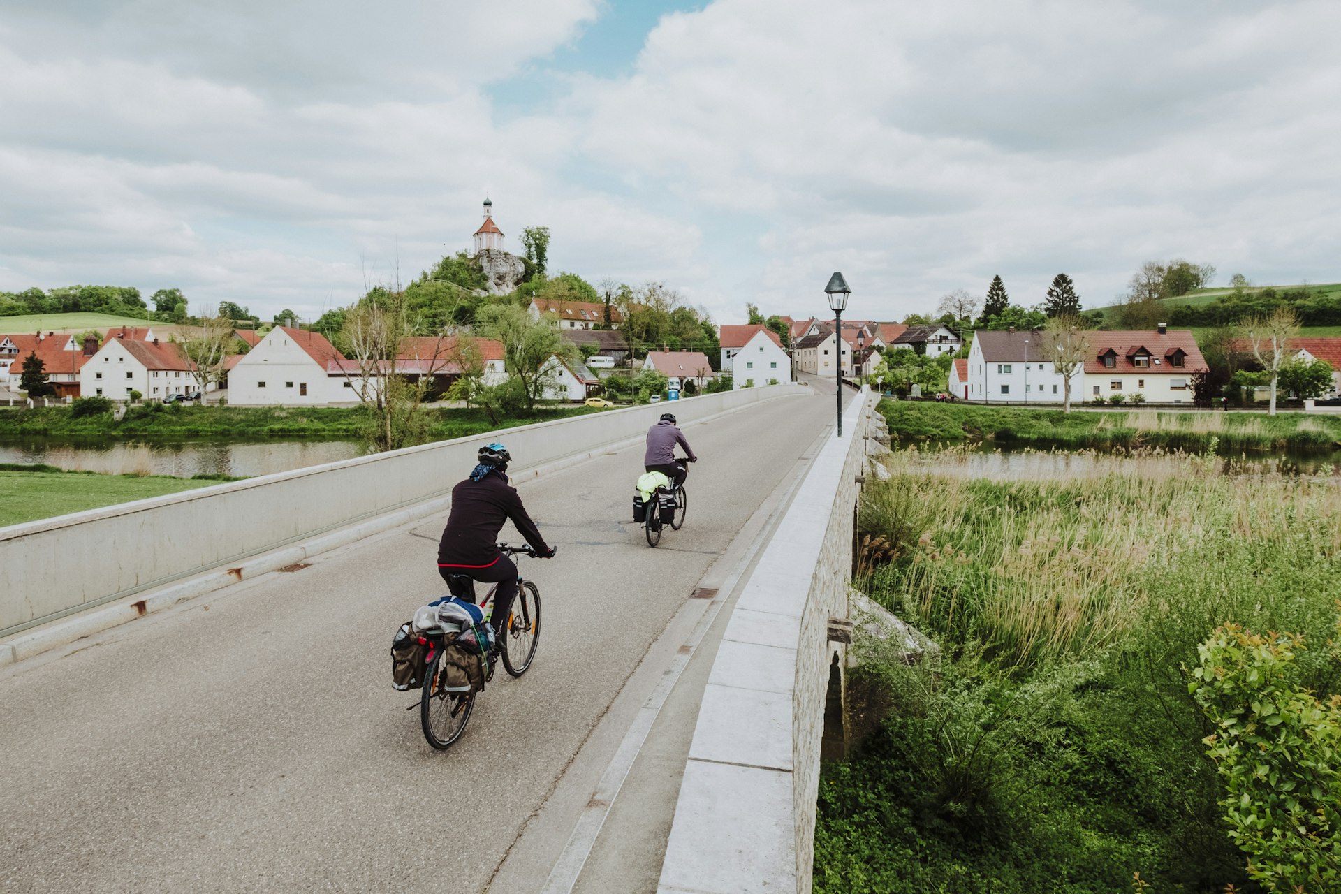 Two cyclists on bikes with luggage ride across a bridge entering a medieval town