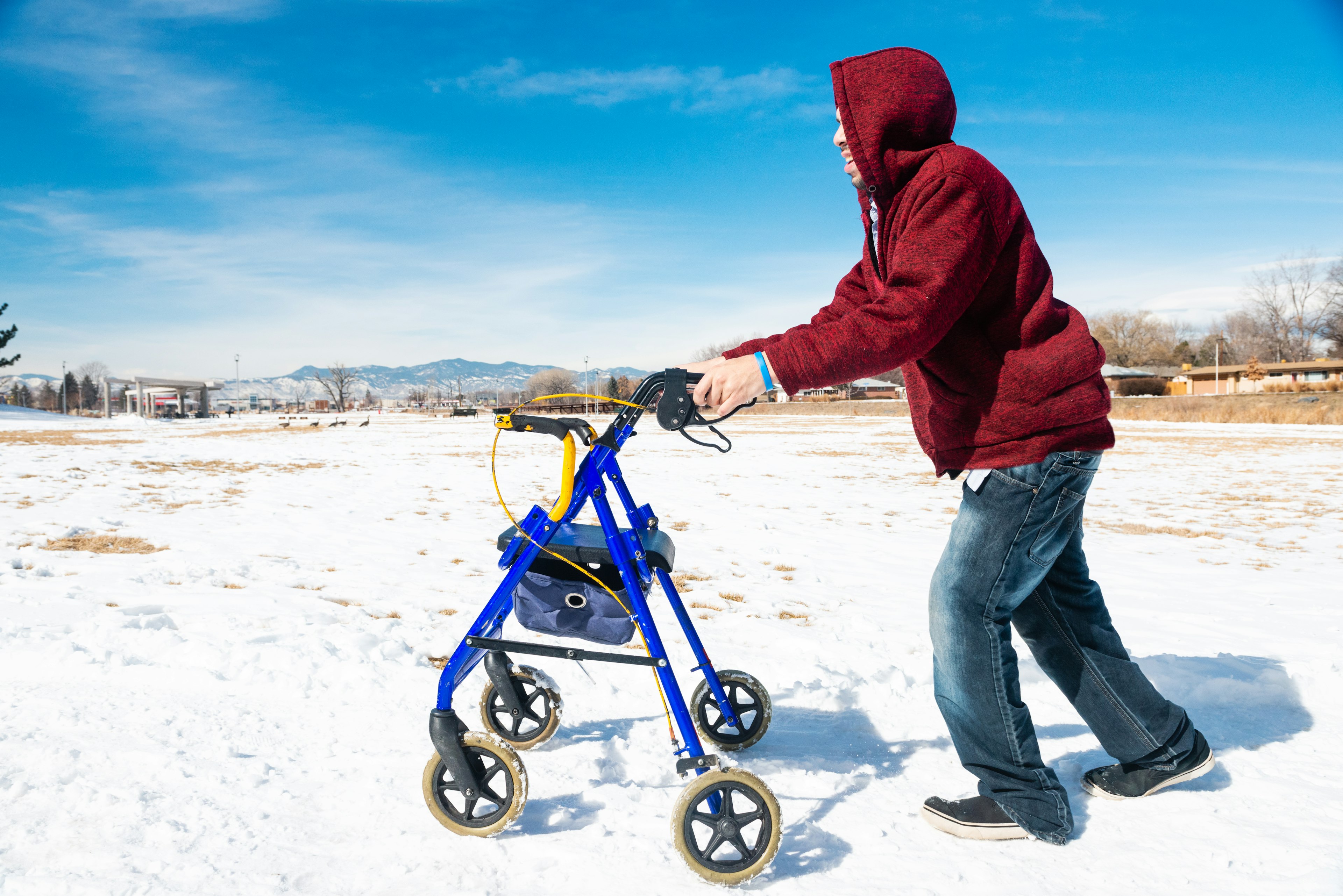 A side view of a young, disabled Hispanic Millennial man using a walker for assistance outdoors to walk along the snow covered park sidewalk in Denver, Colorado on a winter day.