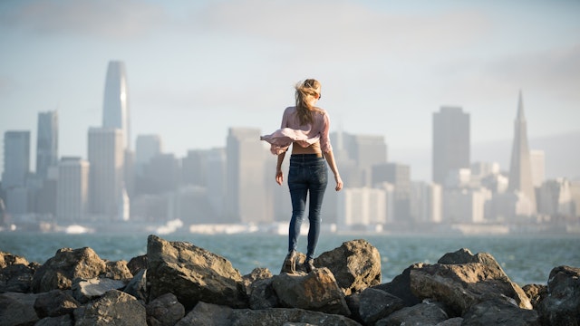 A woman stands on a rocky shore with her back to the camera, looking out at a city skyline across the water.