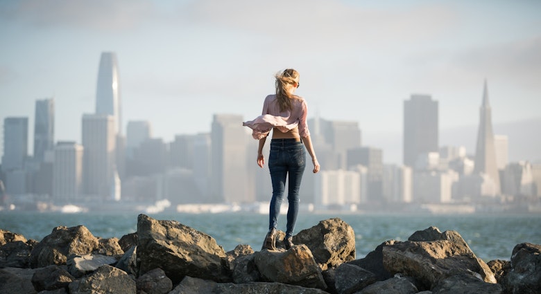 Young woman standing on rocks with the San Francisco skyline in the background.
1164629701