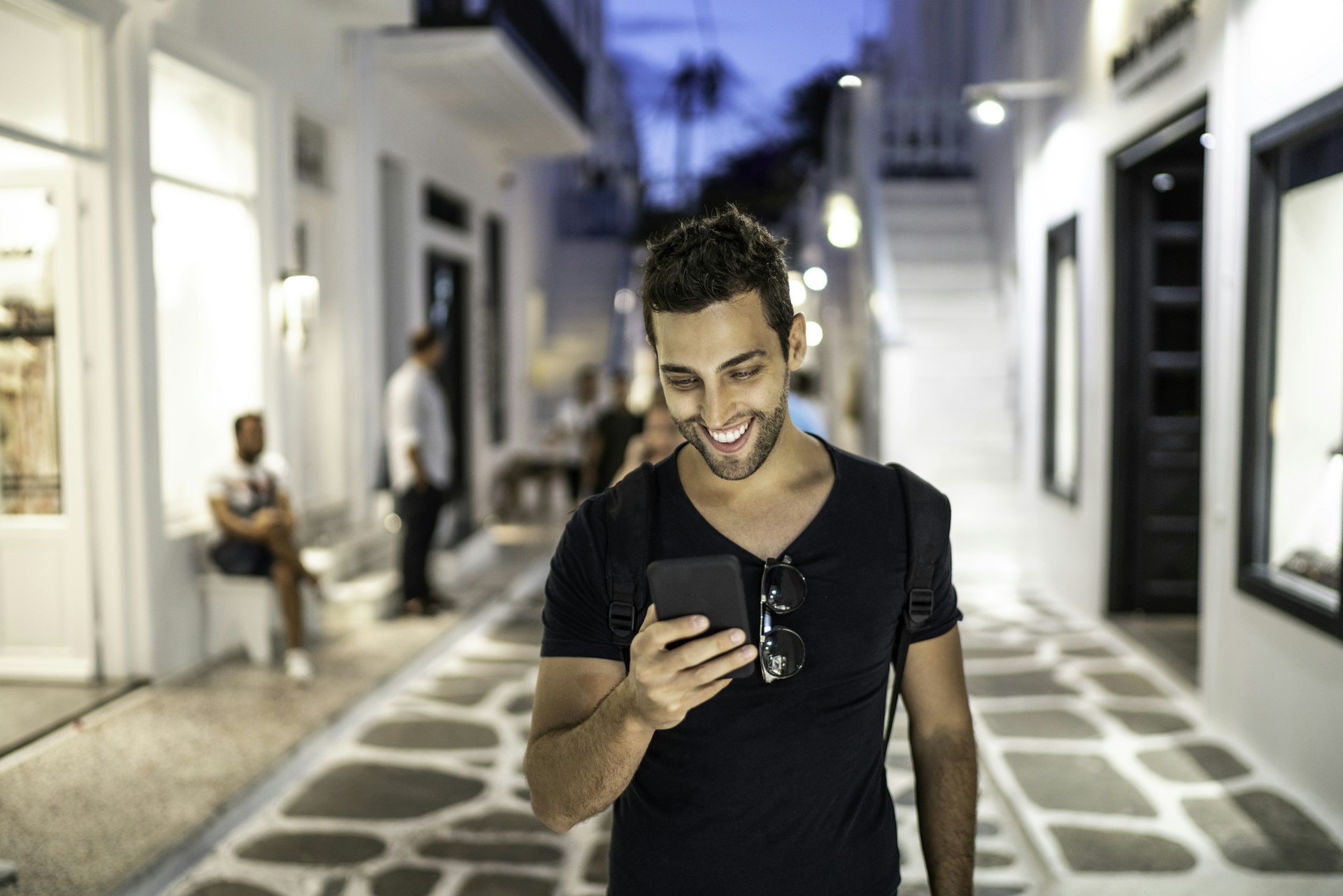A young man smiles as he looks down at his smartphone whilst walking along a greek street