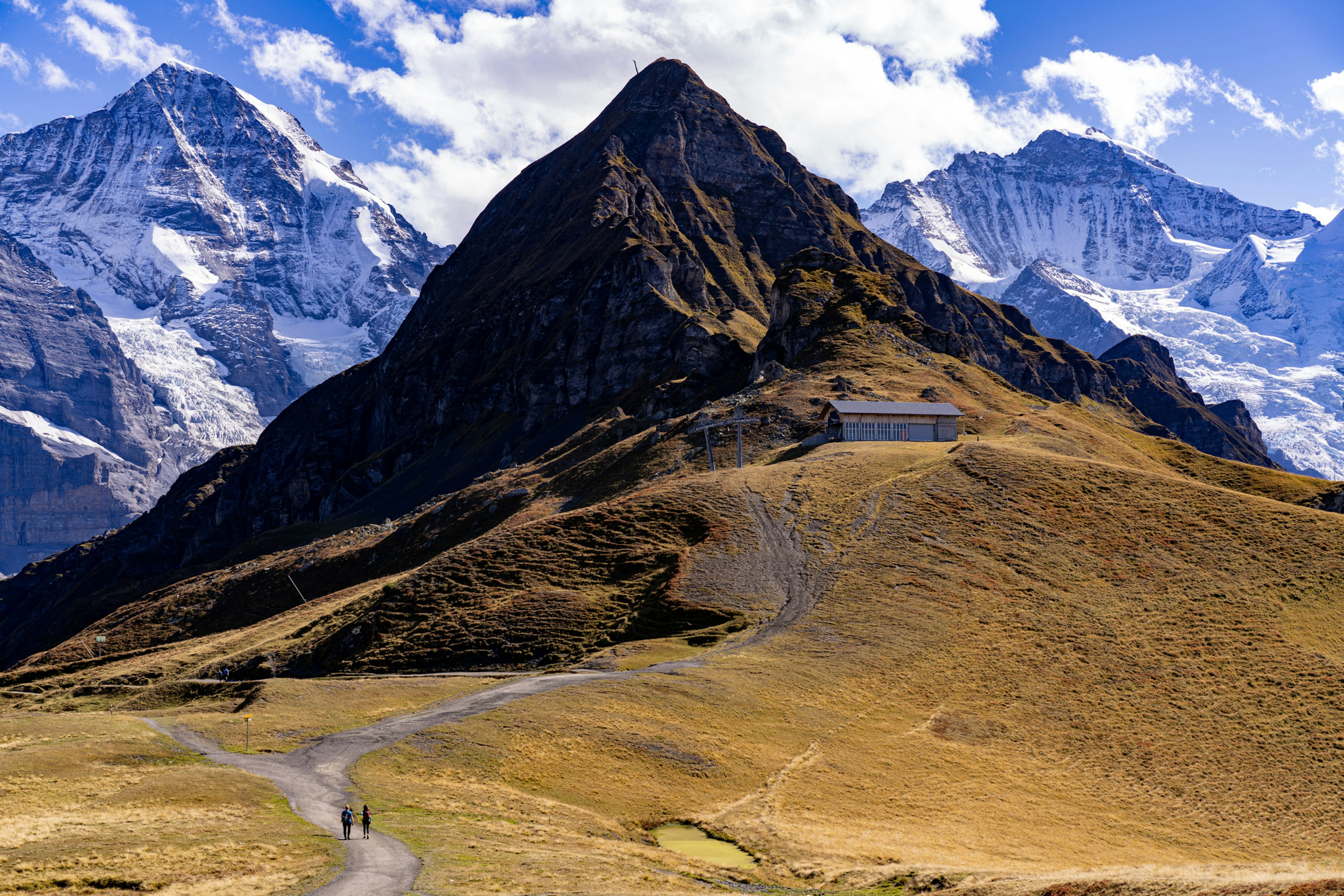 Two hikers walk along a pathway towards a mountain peak