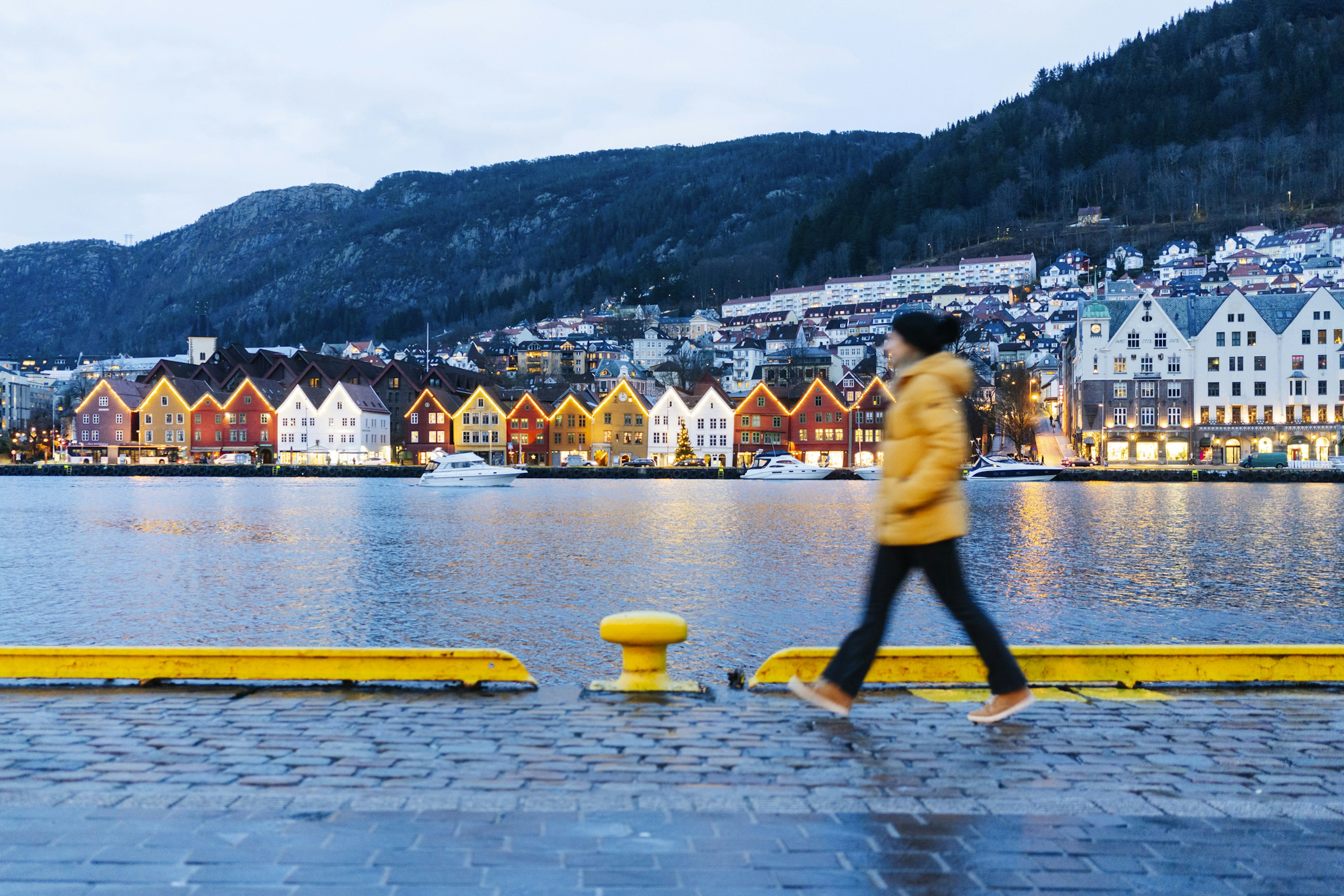 A woman wearing a hat and winter coat walks along a waterfront backed by colorful buildings