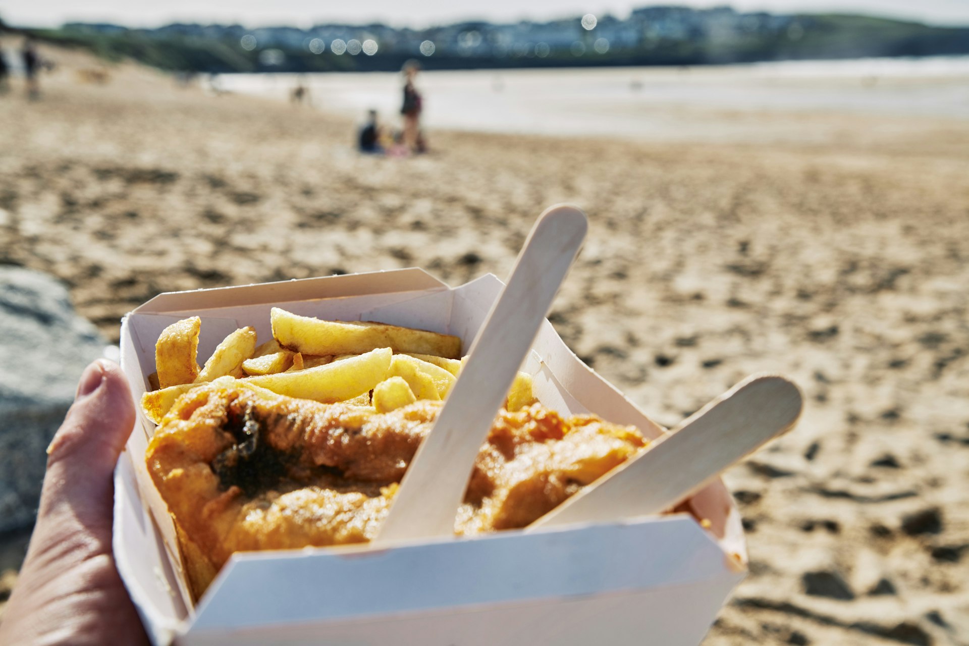 A hand holding a box of fish and chips at the beach on a bright sunny day