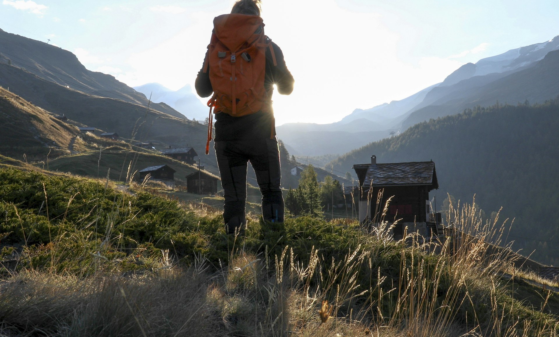 A hiker with a backpack looks off to distant Alps as huts line up in front of him