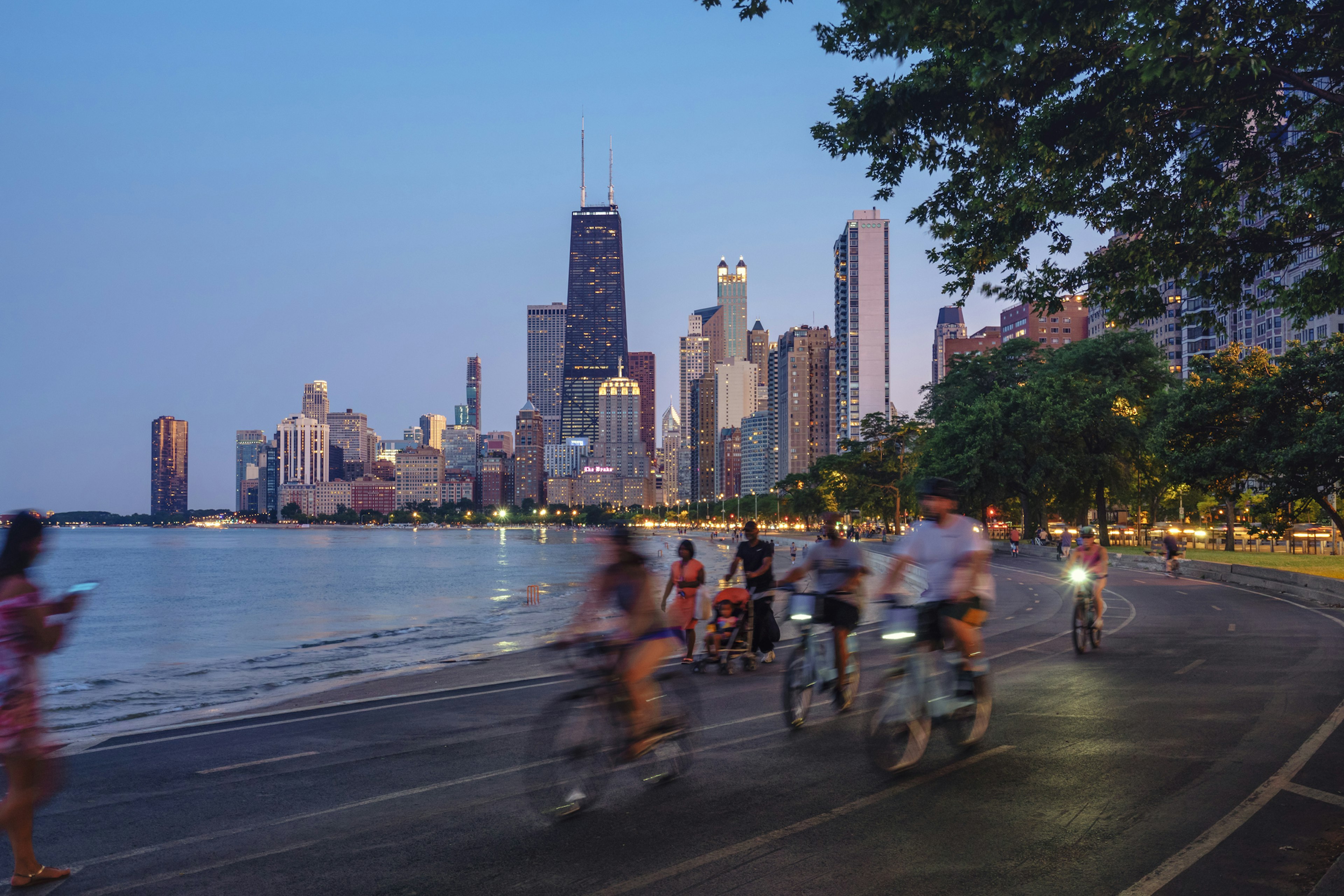 People on bicycles follow a trail beside a lake in a large city. Skycrapers dominate the sky behind