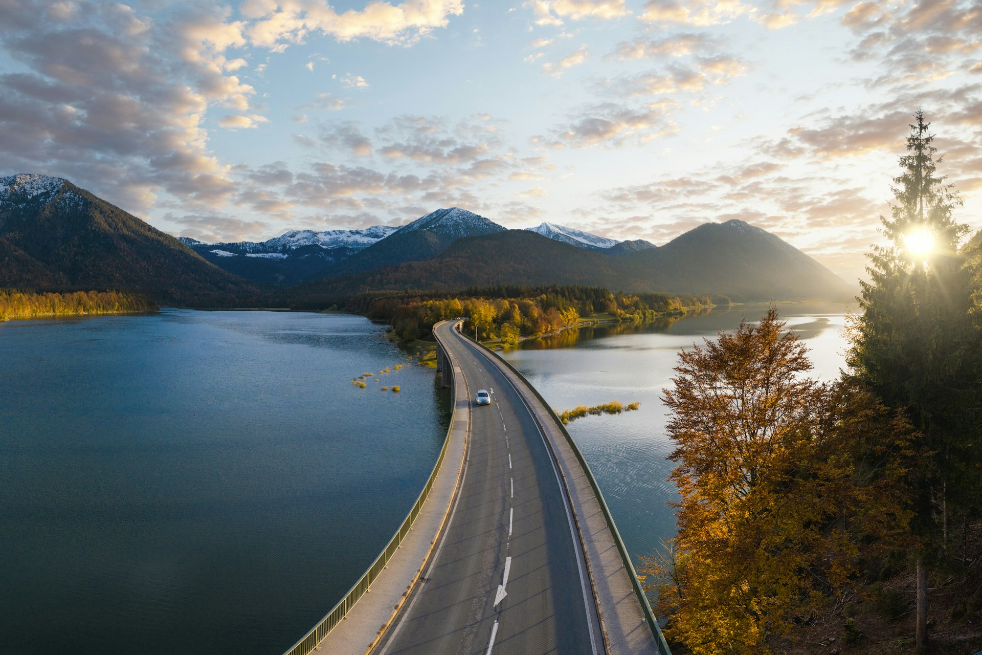 A car follows a well-maintained road over a lake as the sun sets behind distant mountains