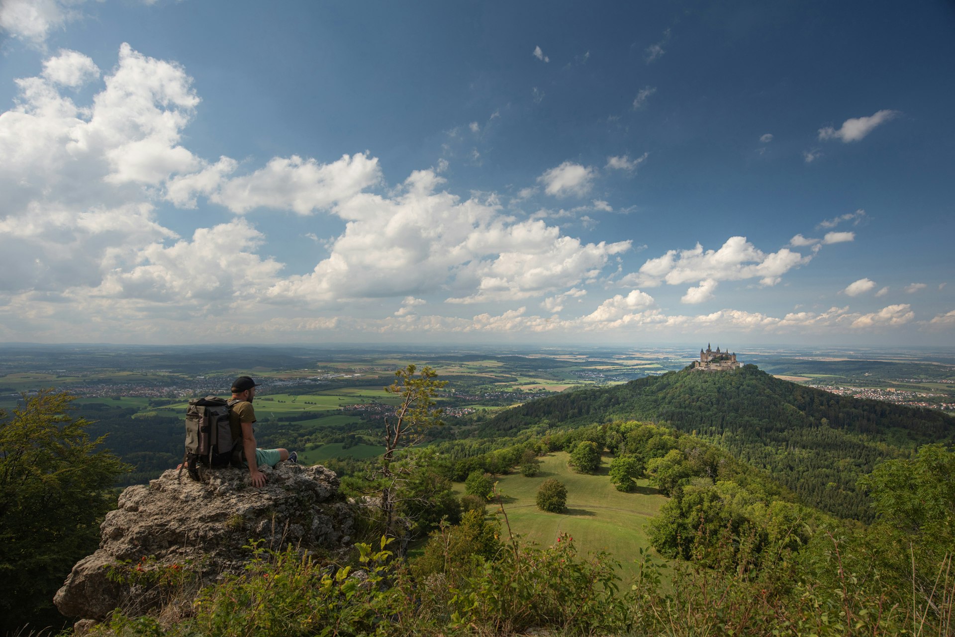 A hiker sits at a viewpoint looking across a green valley towards a castle on a hill