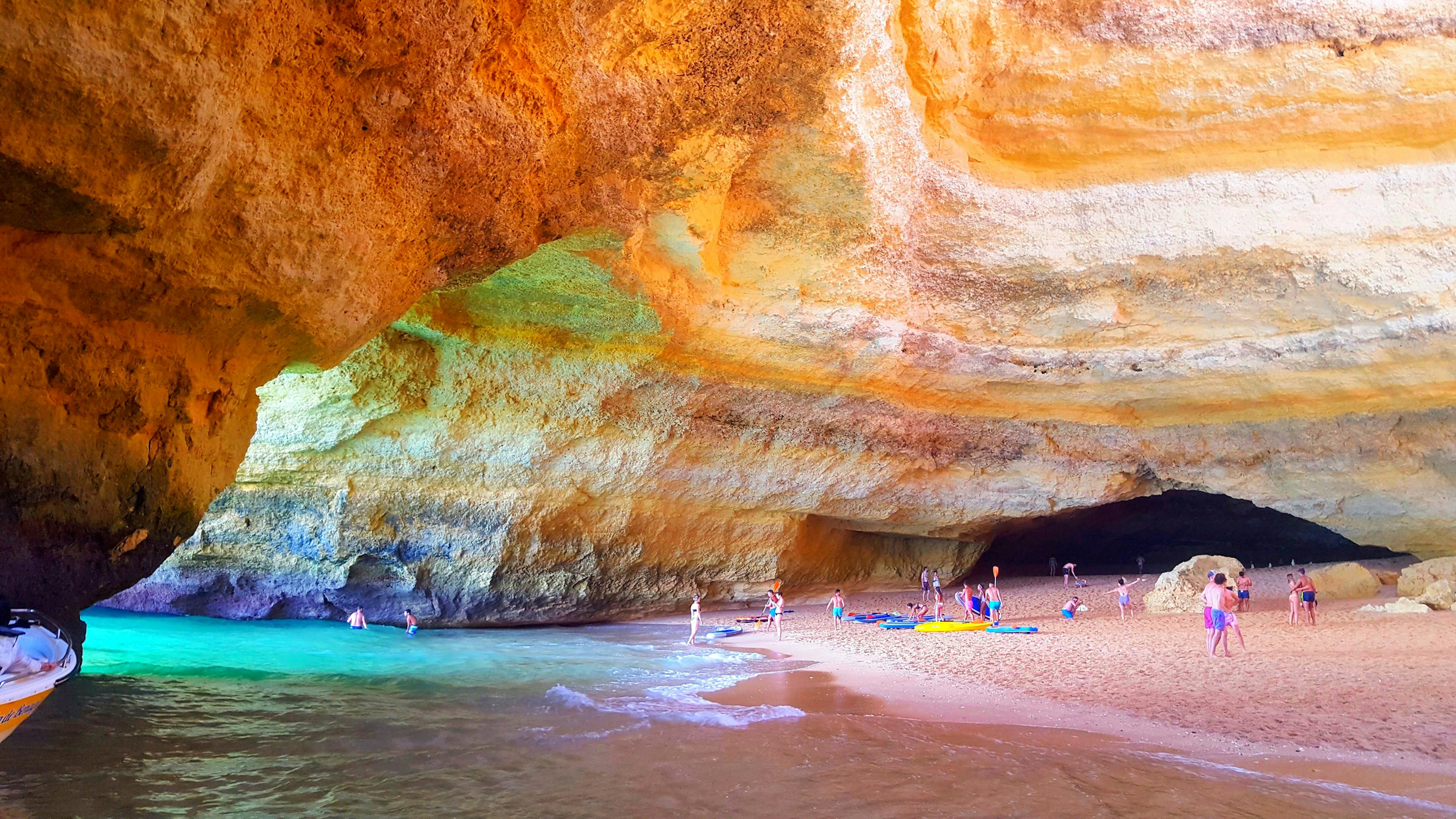 People play on a beach within a vast sandstone cave as the turquoise water laps the sand