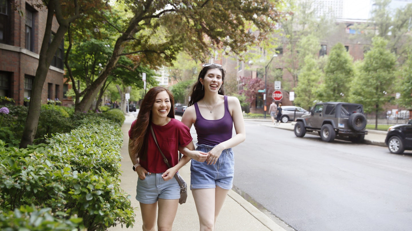 Two young Generation Z women, with long dark hair, laugh while walking along the street, while wearing casual summer clothing.
1369762415
Two women walking arm in arm down a Chicago street