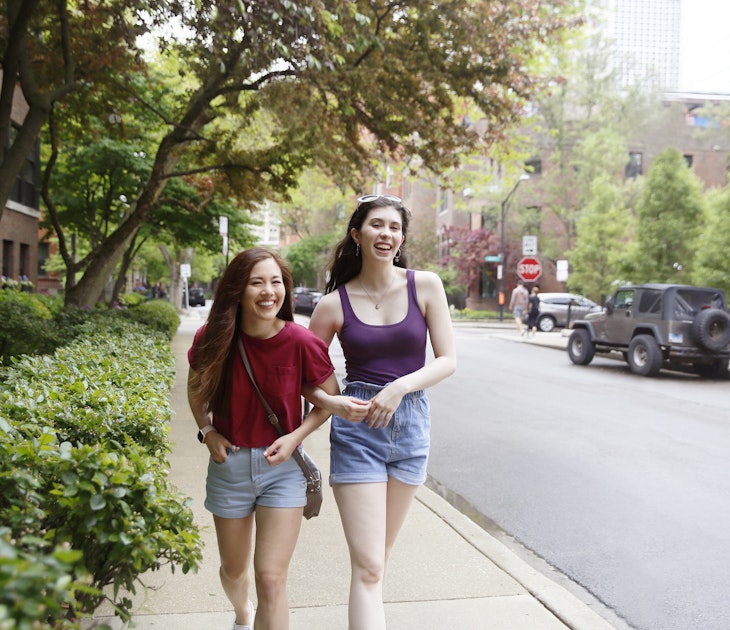 Two young Generation Z women, with long dark hair, laugh while walking along the street, while wearing casual summer clothing.
1369762415
Two women walking arm in arm down a Chicago street
