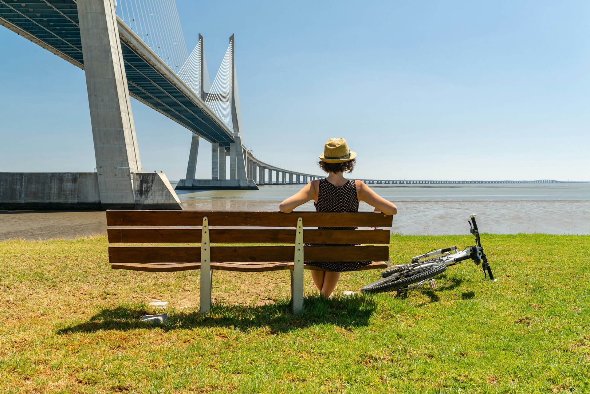 A woman with a bike rests on a riverside bench gazing out towards a huge bridge 