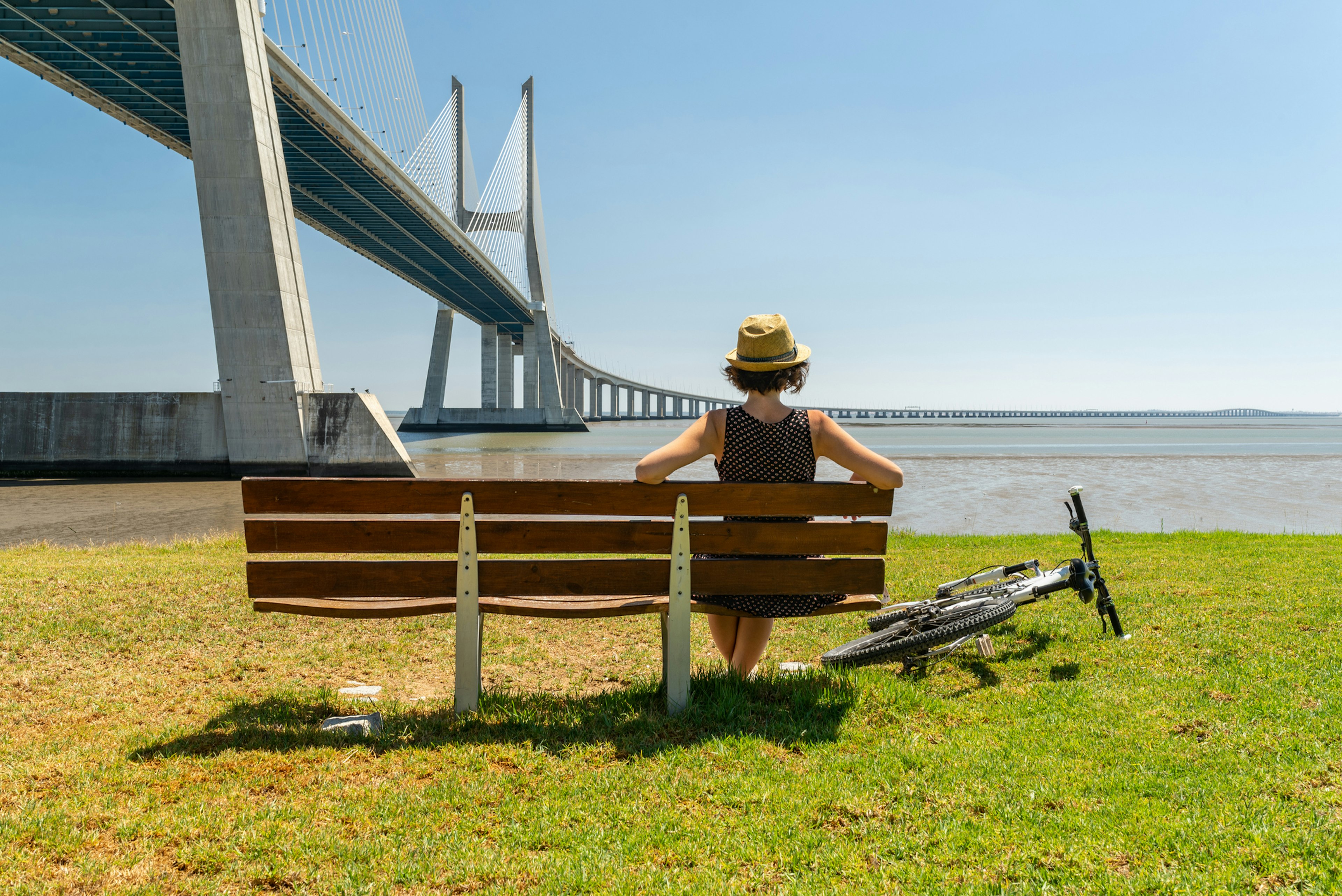 A woman with a bike rests on a riverside bench gazing out towards a huge bridge